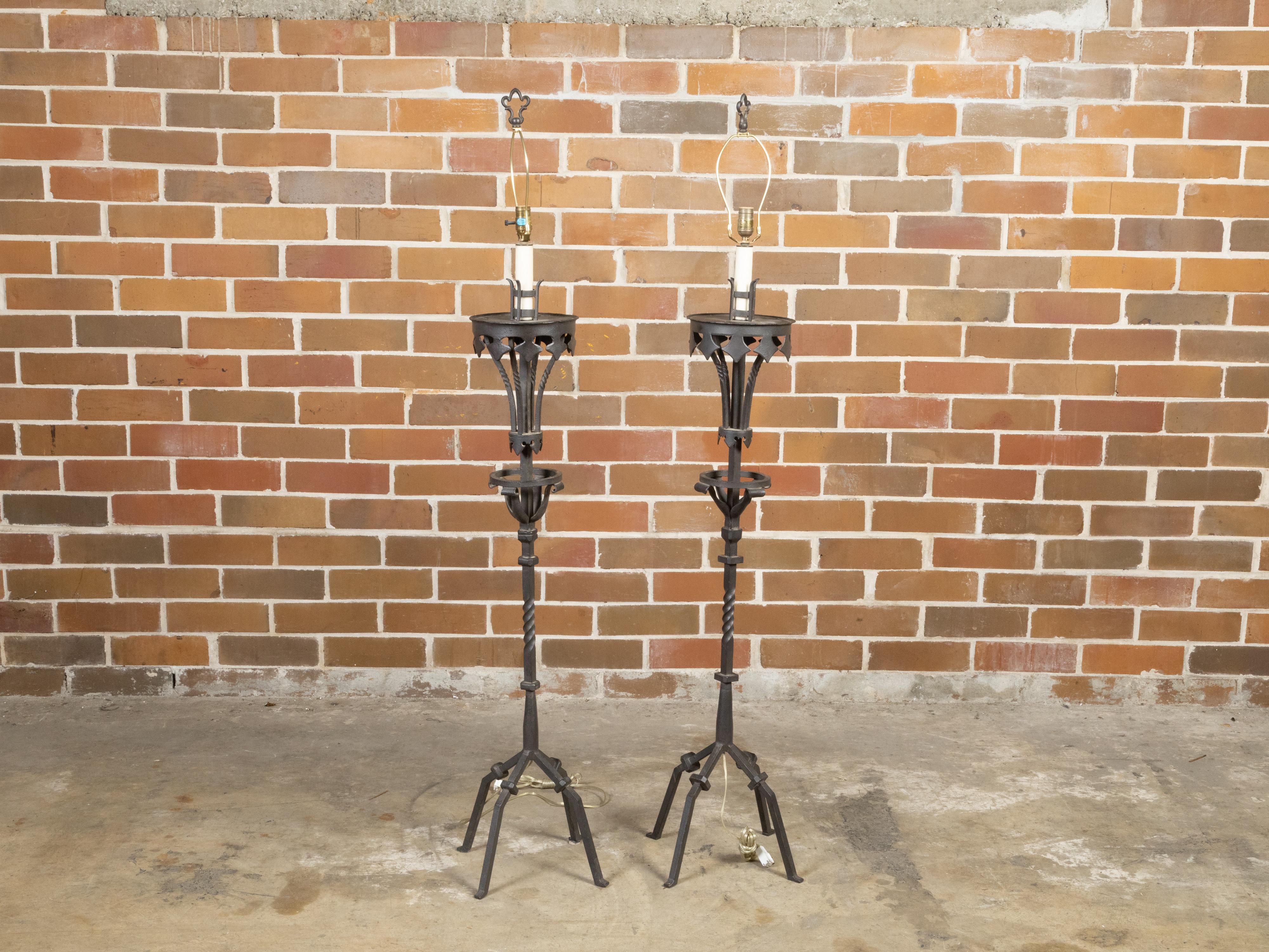 A pair of French Gothic style iron torchères floor lamps from the mid 20th century, with single lights, in-curving diamond motifs, twisted accents and quadripod bases. Created in France during the Midcentury period, this pair of Gothic style iron