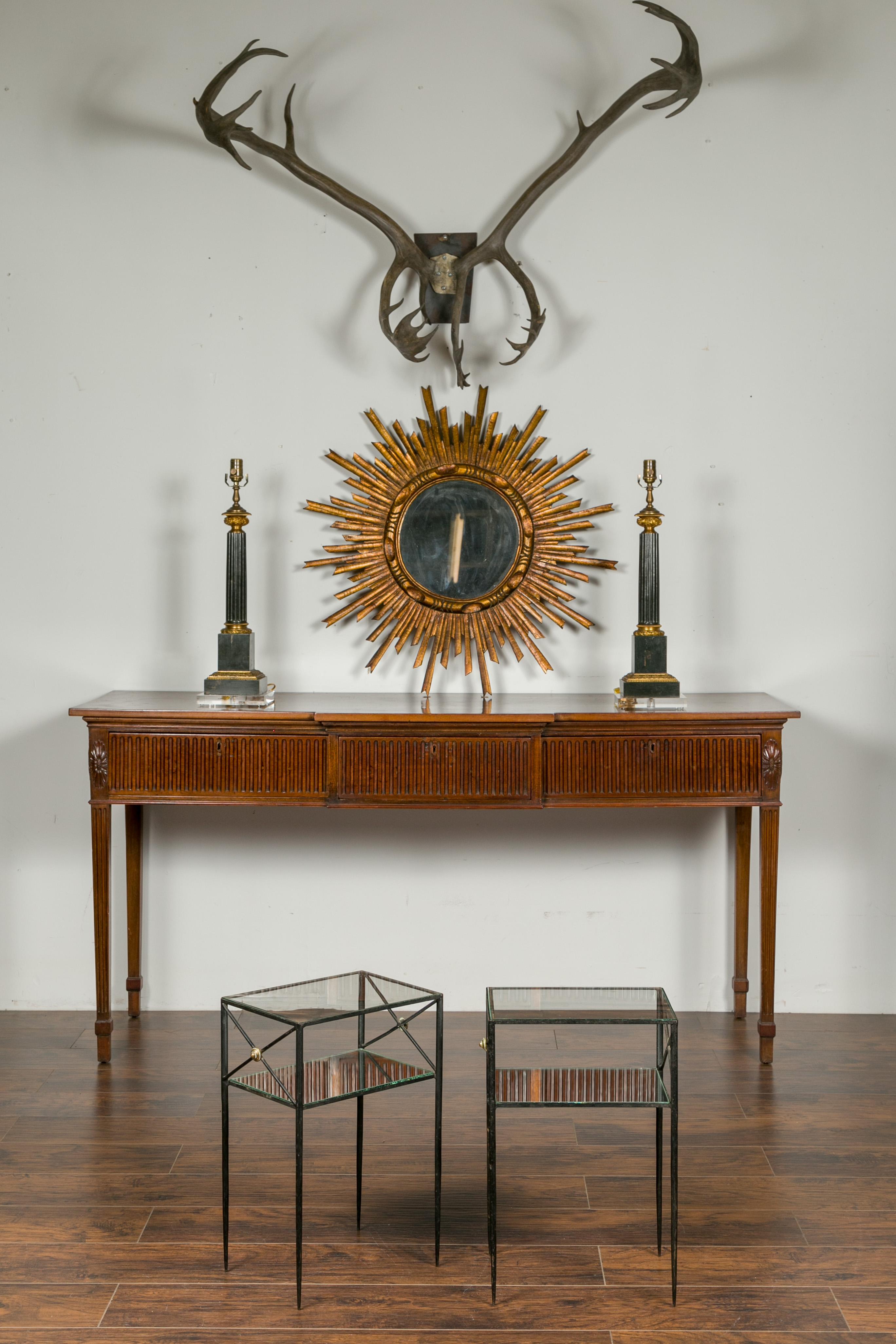 A pair of French vintage iron side tables from the mid-20th century, with brass accents, glass tops and mirrored shelves. Born in France during the midcentury period, each of this pair of drinks tables features an iron structure securing a glass top