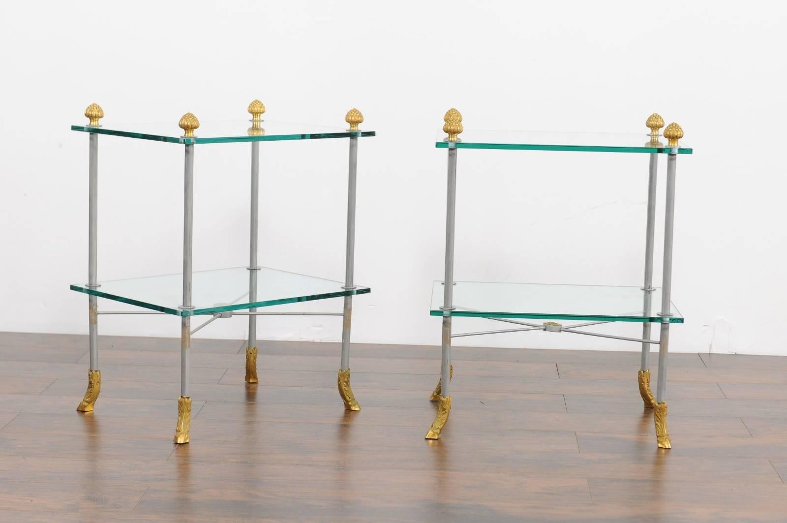 Pair of French vintage steel and brass tiered side tables from the mid-20th century with glass shelves and hoofed feet. Each of this pair of French side tables features a simple steel structure made of four straight legs, accented with brass