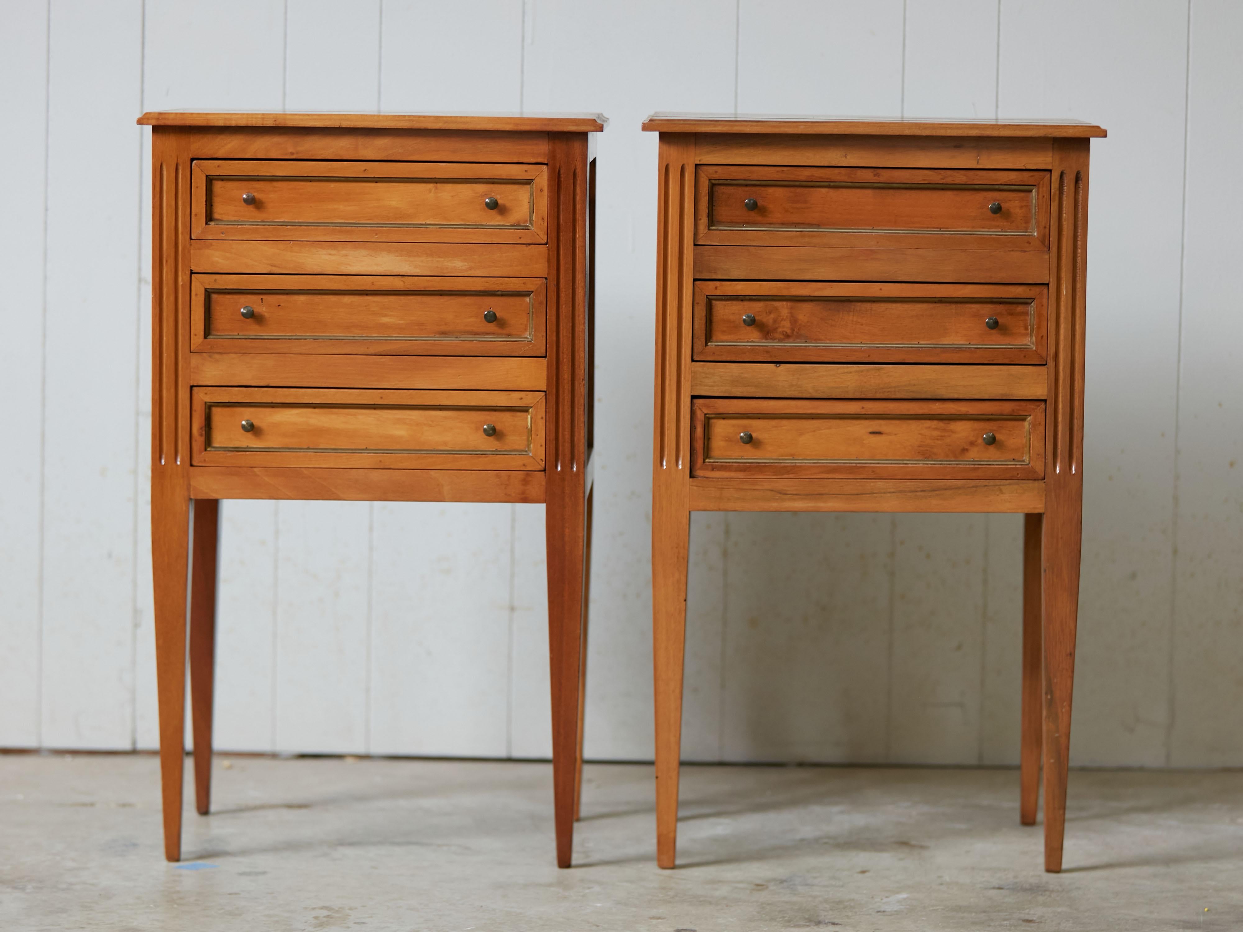 A pair of French walnut bedside tables from the mid 20th century, with three drawers, gilded frames and fluted motifs. Created in France during the Midcentury period, each of this pair of tables features a rectangular planked top sitting above three