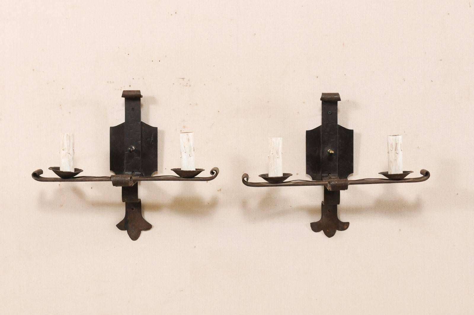 A pair of French two-light iron sconces from the mid-20th century. These vintage French hand-forged sconces each feature a horizontally placed bar, with delicately scrolled ends, which provide support to the two iron bobèches and painted candle