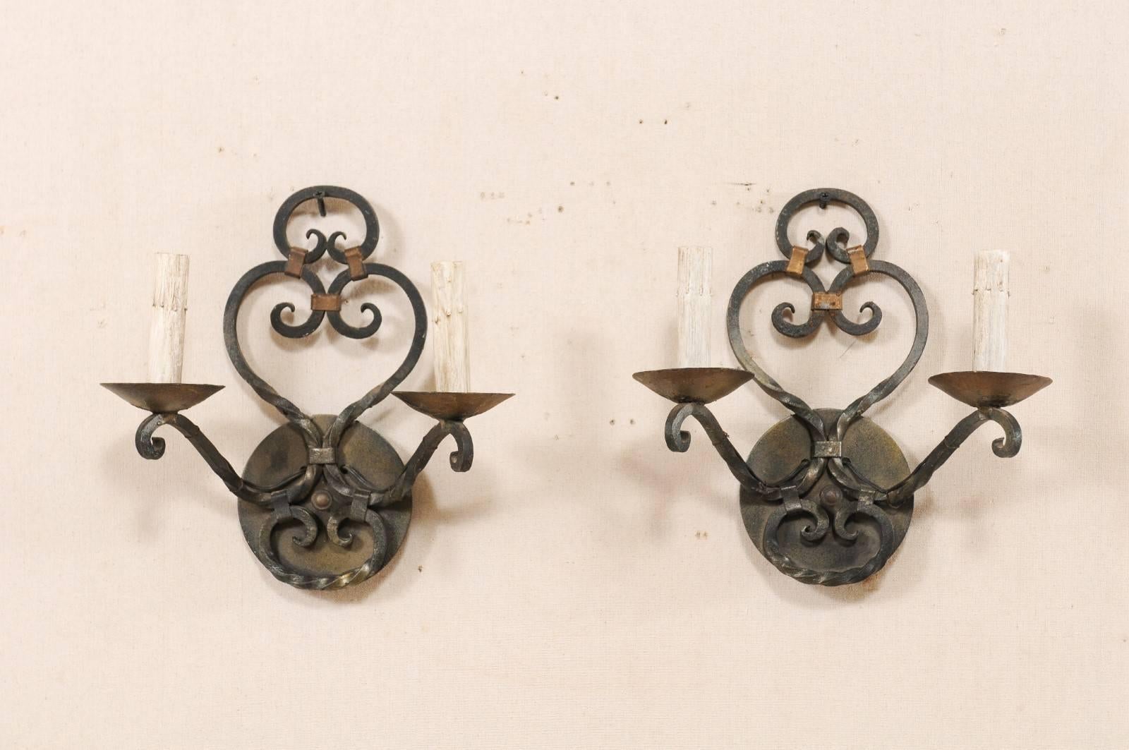 A pair of French midcentury two-light scrolled iron sconces. These vintage French hand-forged iron sconces each feature two scrolled arms, which delicately lift out and away from the round-shaped back plate, supporting their iron bobèches and