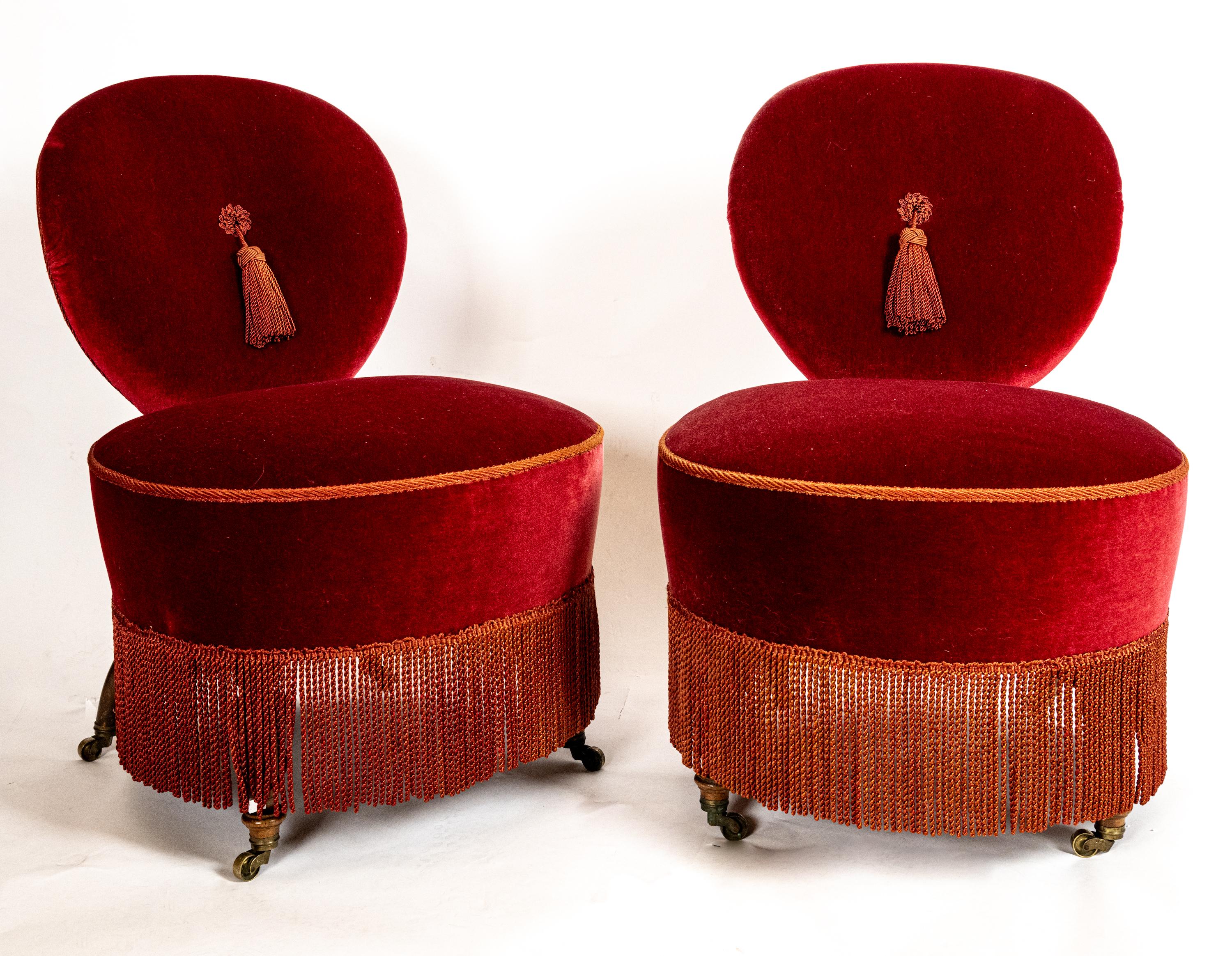 A pair of French Napoleon III period Fez slipper chairs on brass castors with the seat and back fully upholstered in a rich crimson velvet. Having a large passementerie tassel tufted at the center of the front, vermilion piping along the edges, and
