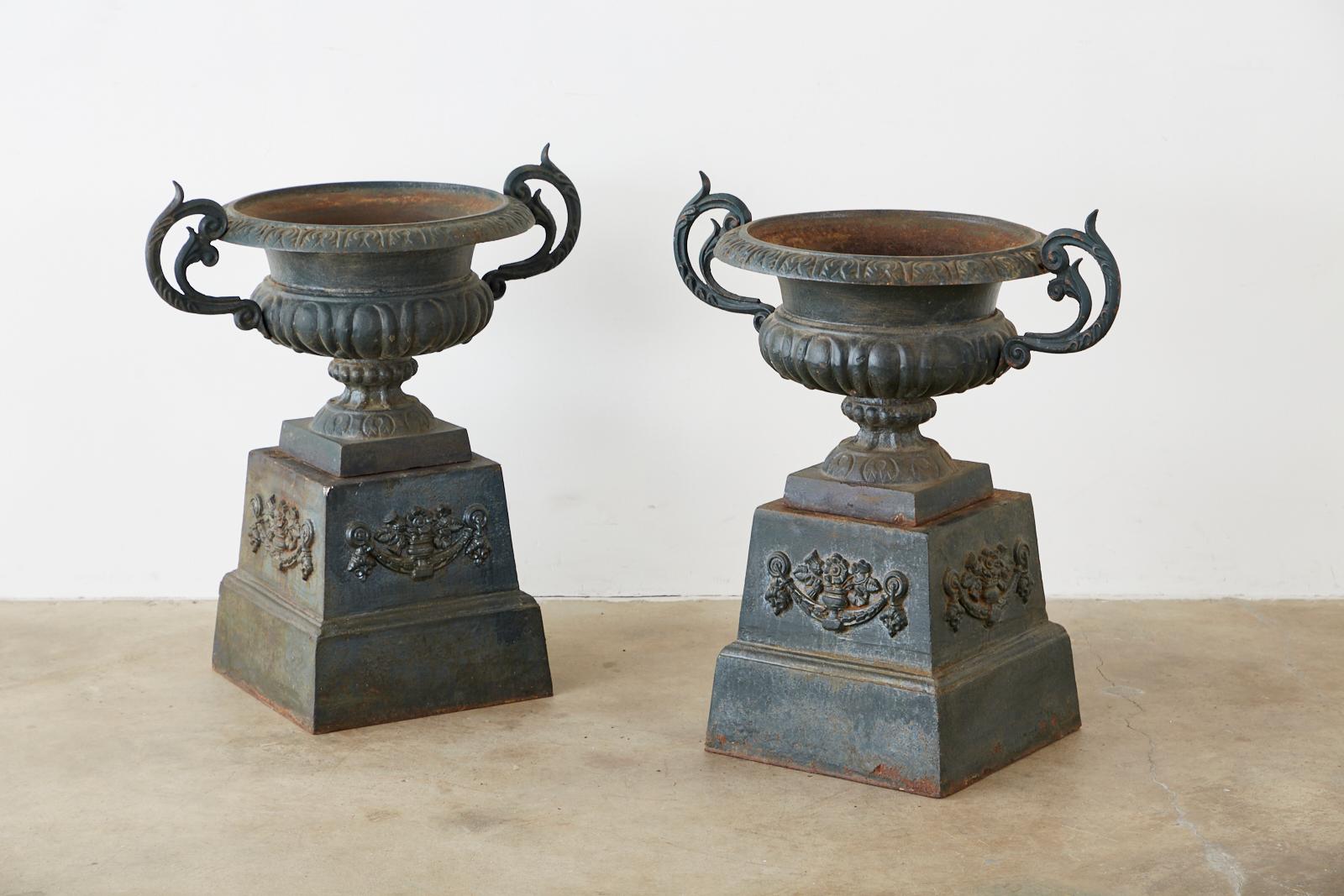 Large pair of 19th century French cast iron garden urns or jardinière planters on pedestals. The jardinières feature large scrolled handles on each side. Each urn sits atop a stepped pedestal base decorated with floral and acanthus swag in the