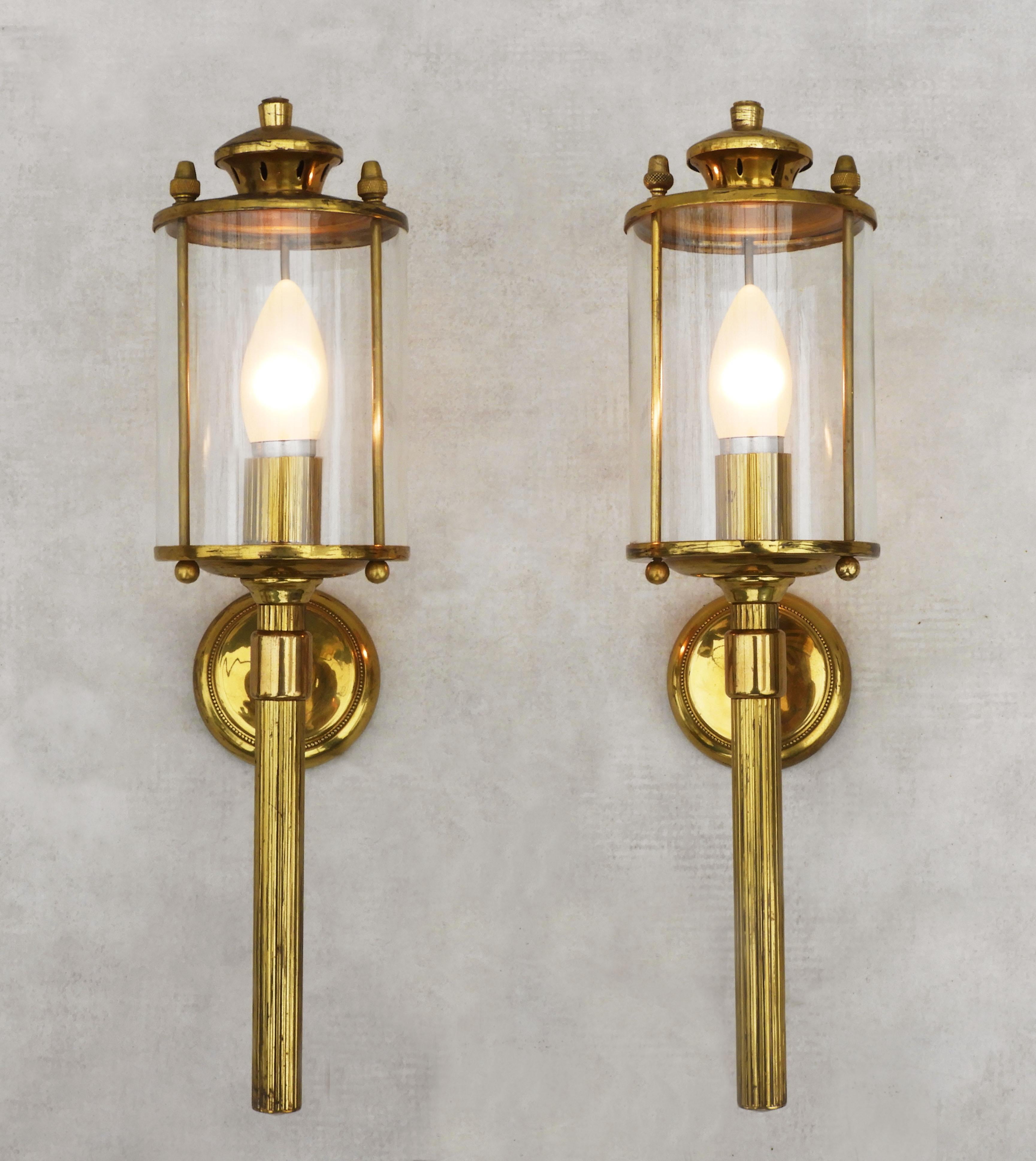 Great pair of Jansen-style brass wall lanterns from mid-century France.  A duo of torchère style lamps with cylindrical glass shades, neoclassical detailing and acorn-shaped finials.  A stylish, French, sophisticated modern take on a timeless