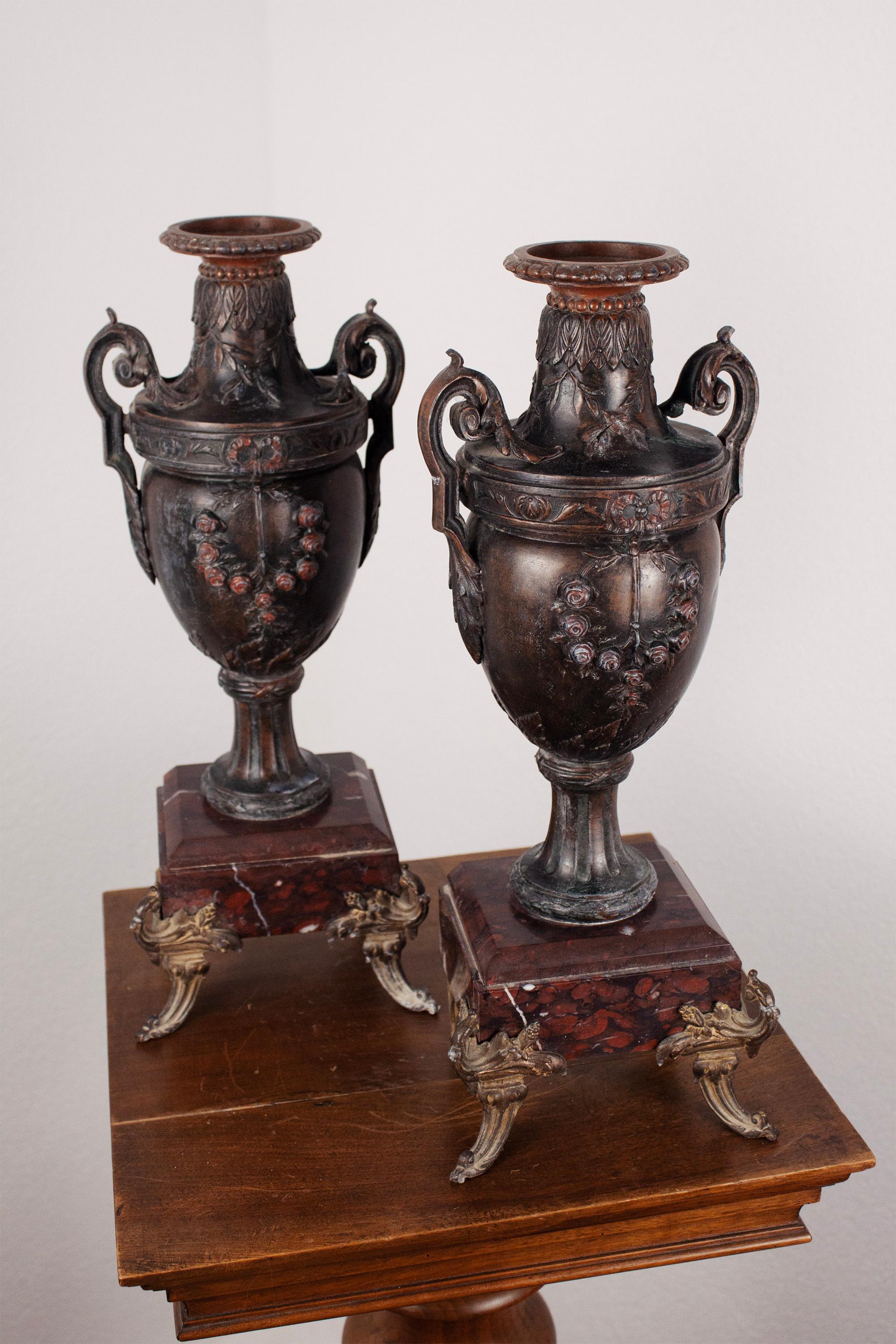 This elegant pair of French Neoclassical urns features finely cast bronze bodies decorated with floral garlands and classical motifs. Each urn rests on a red marble plinth with ornate gilt bronze mounts and scrolled feet, combining rich textures and
