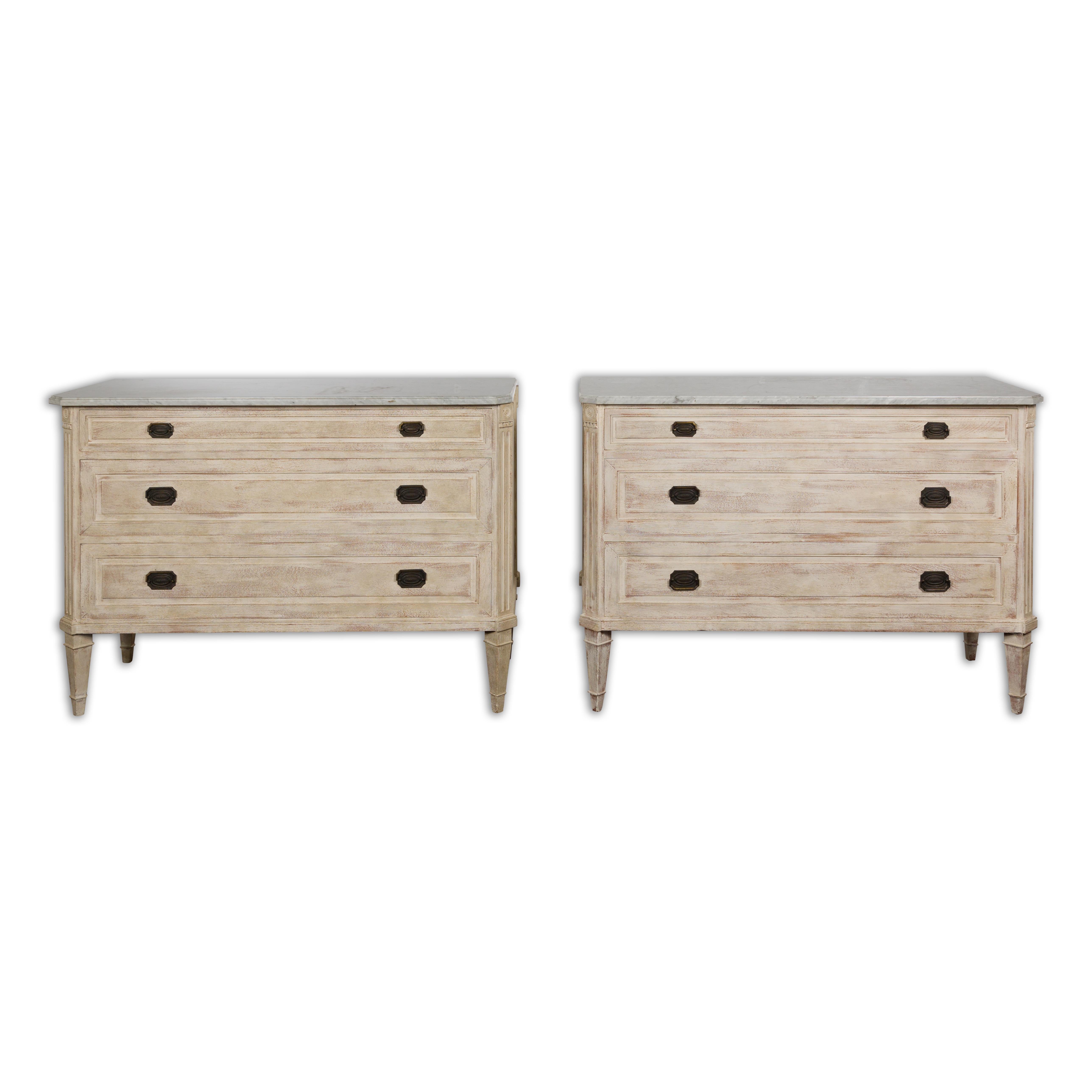 A pair of French Neoclassical style painted wood three-drawer chests from circa 1930 with light grey marble tops, graduated drawers, carved fluted side posts and tapered legs. Elevate your living space with this captivating pair of French