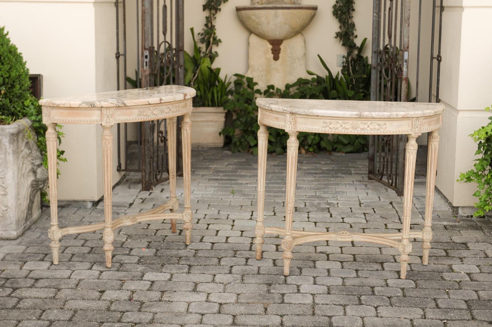 A pair of French neoclassical style carved and painted wooden demi-lune console tables from the early 20th century, with marble tops and fluted legs. Born in France during the Belle Époque era, each of this pair of demilunes features a semi-circular