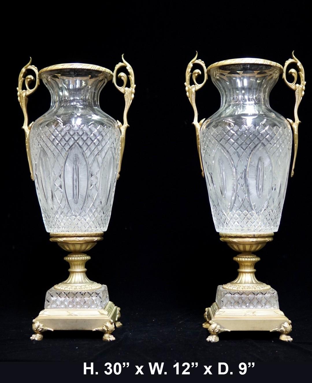 Impressive and large pair of French Louis XVI style ormolu and cut crystal urns,
mid-20th century.  

Each urn is centered by an ovoid shape cut-crystal body flanked by two ormolu handles, over a gilt bronze reeded foot, above a conforming cut