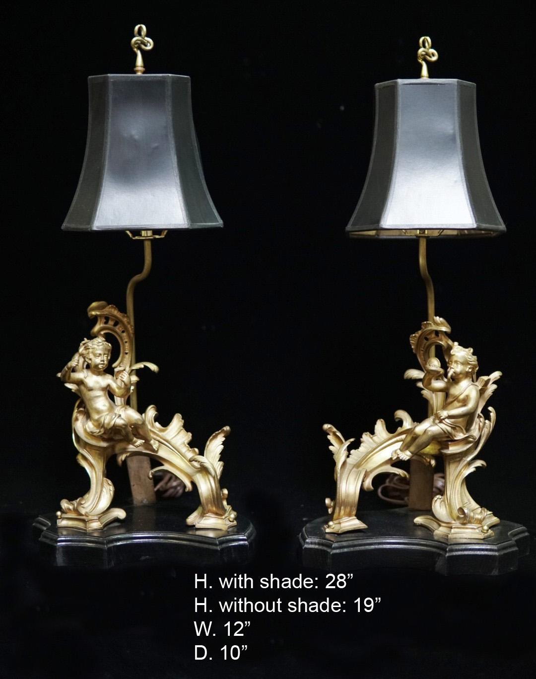 Pair of French ormolu chenet mounted lamps with black rectangular shades,
Late 19th century.
Each lamp is mounted with a ormolu acanthus motif chenet with a putti, raised on a conforming stepped black base. 
Shades not