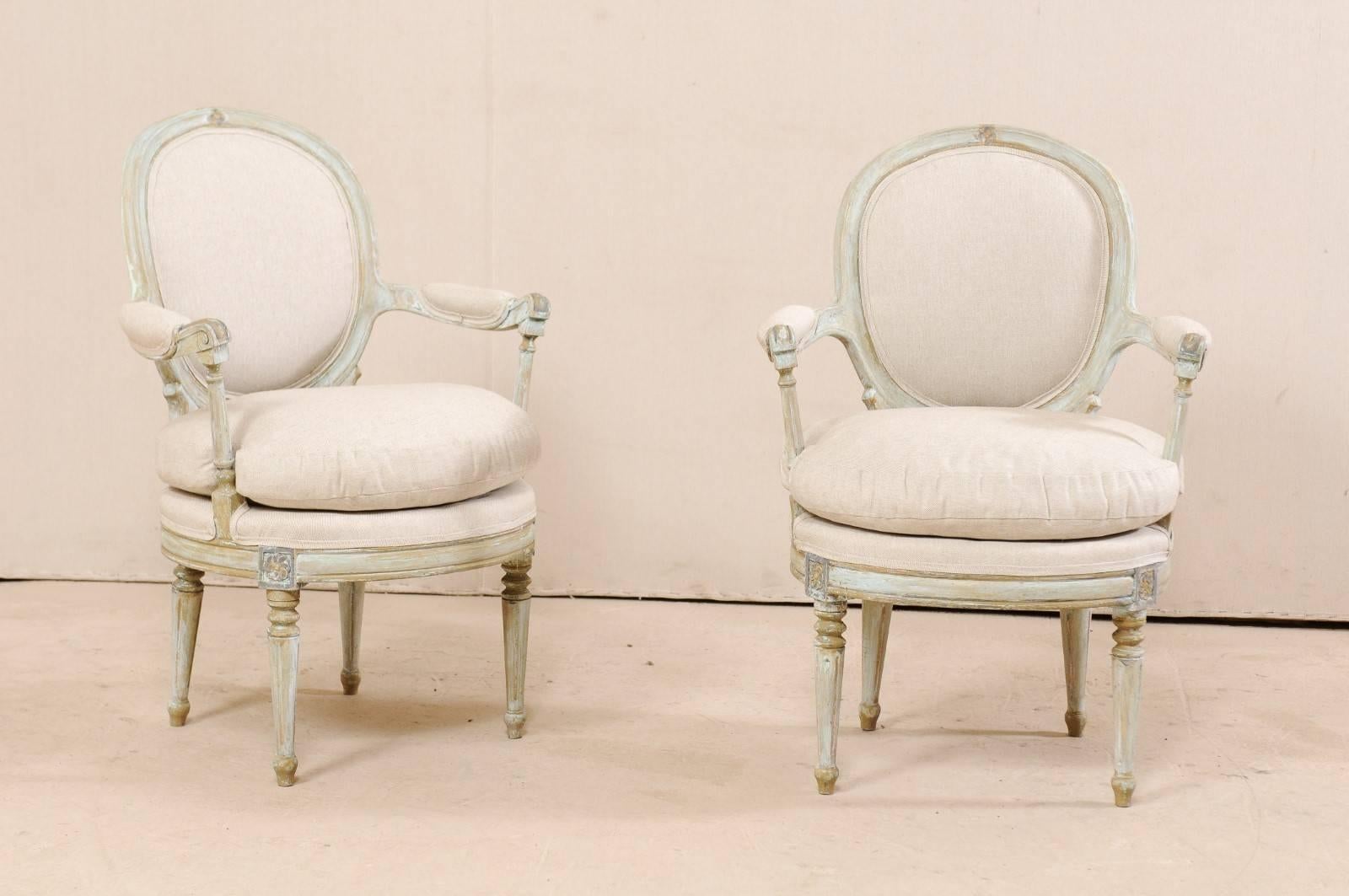 A pair of French painted wood and upholstered bergère chairs. This elegant pair of French armchairs each feature oval-shaped upholstered backs with a delicately carved flower at top center rail. There are manchette padded arms with carved curled