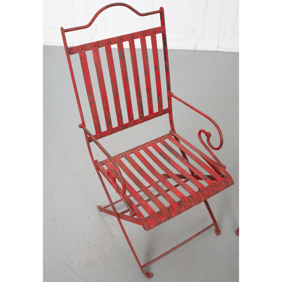 This pair of French painted red antique iron garden chairs has a playful design. They have an arched back of vertical slats and seat with artfully scrolled arms. The legs cross and are connected by stretchers. They would make a whimsical addition to