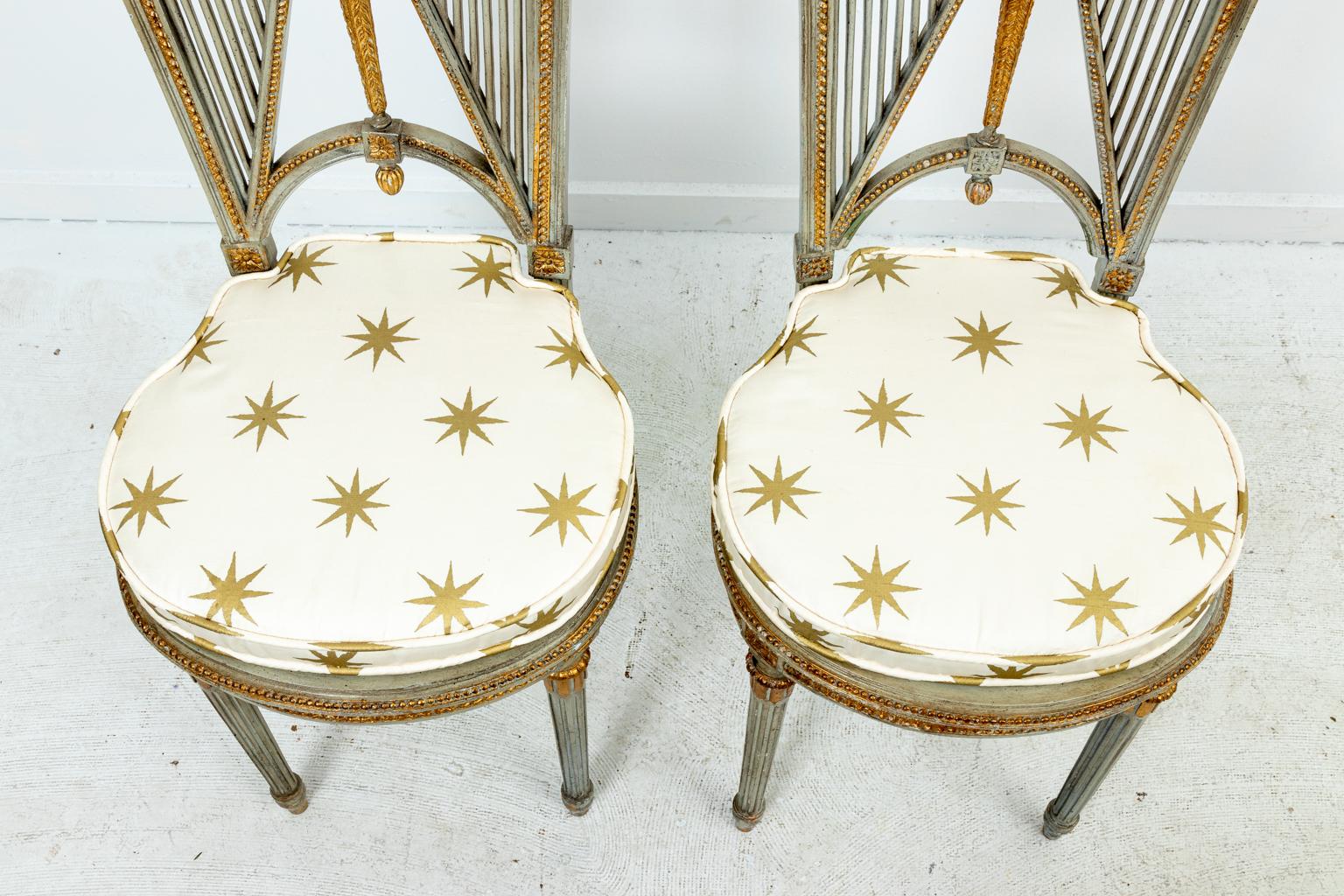 Pair of French painted neoclassical or Empire style side chairs with upholstered starburst seats and gold painted accents. The chairs feature neoclassical motifs such as carved harps on the seat back, rosettes, and beaded trim on the seat base. The