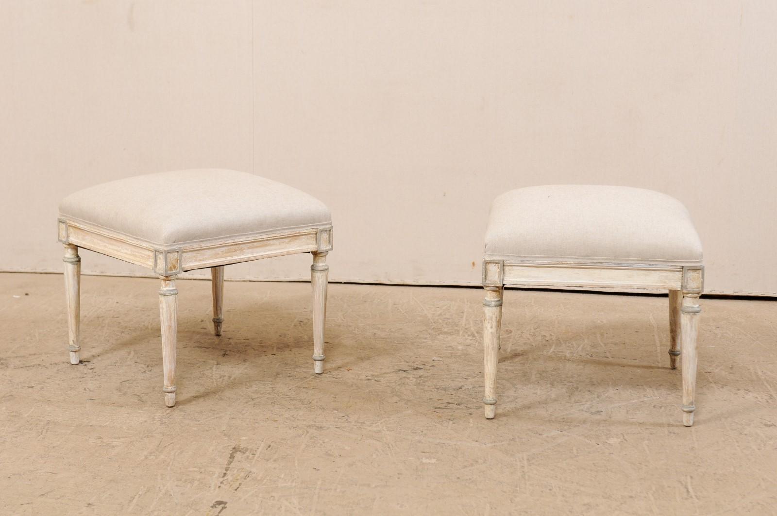 A pair of French wooden stools with upholstered seats from the early 20th century. This antique pair of French stools each feature straight skirts, carved rectangular outline shapes atop each knee, and are presented upon four rounded and gently