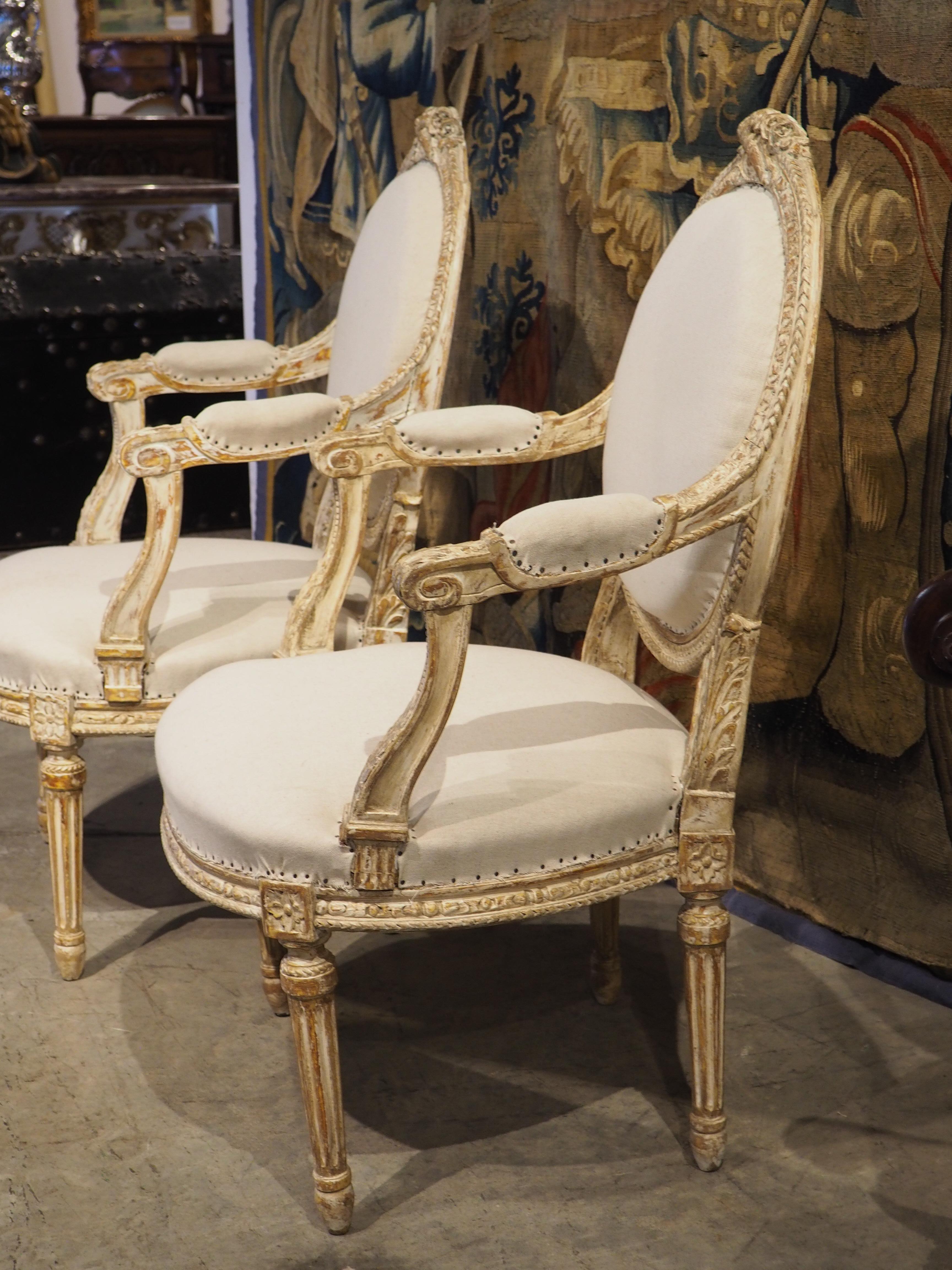 Datant d'environ 1885, cette paire de fauteuils cabriolet à peinture parcellaire sculptée à la main est de style Louis XVI. Les épais médaillons ovales (qui ont conservé leur toile à carreaux d'origine au verso) ont été ornés d'un trophée au niveau