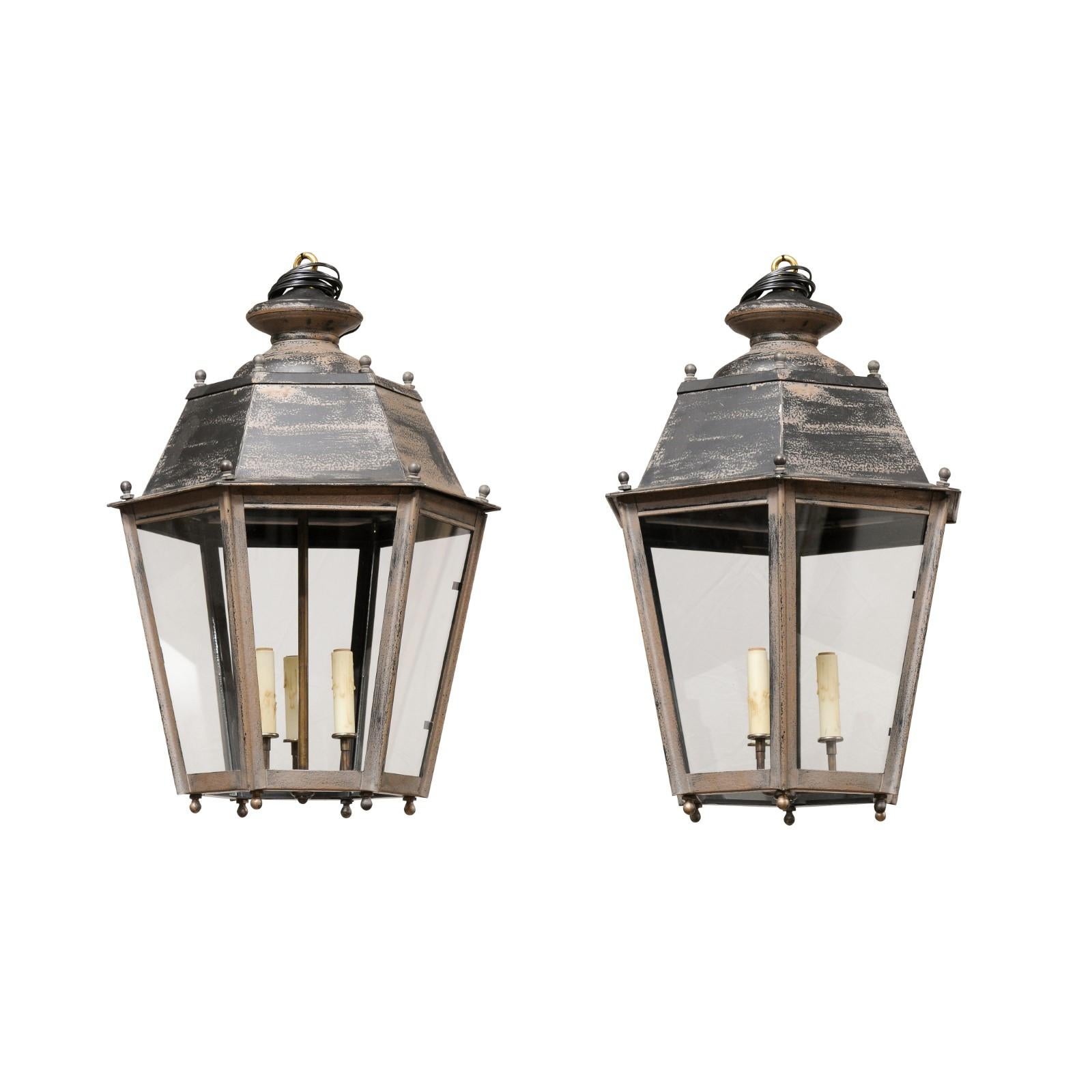 A pair of French patinated metal hexagonal lanterns from the 20th century, with three lights, petite spheres, glass panels and nicely weathered patina. Created in France during the 20th century, each of this pair of patinated metal lanterns features