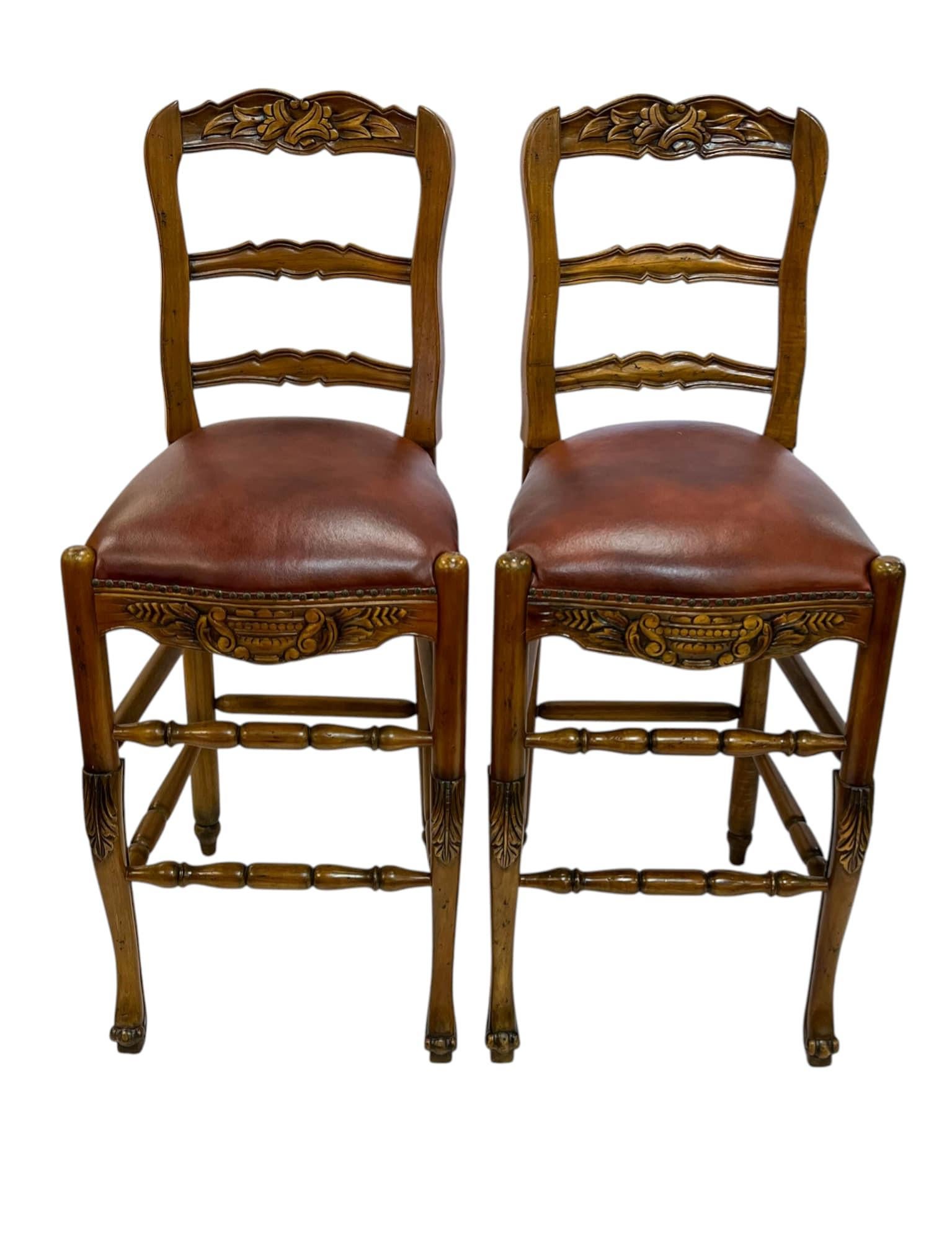 This pair of carved pine bar stools combines the elegance of French Provincial design with the bold details of Jacobean Revival. Each stool features a classic ladder-back with undulating slats and a carved crest, paired with turned stretchers and an