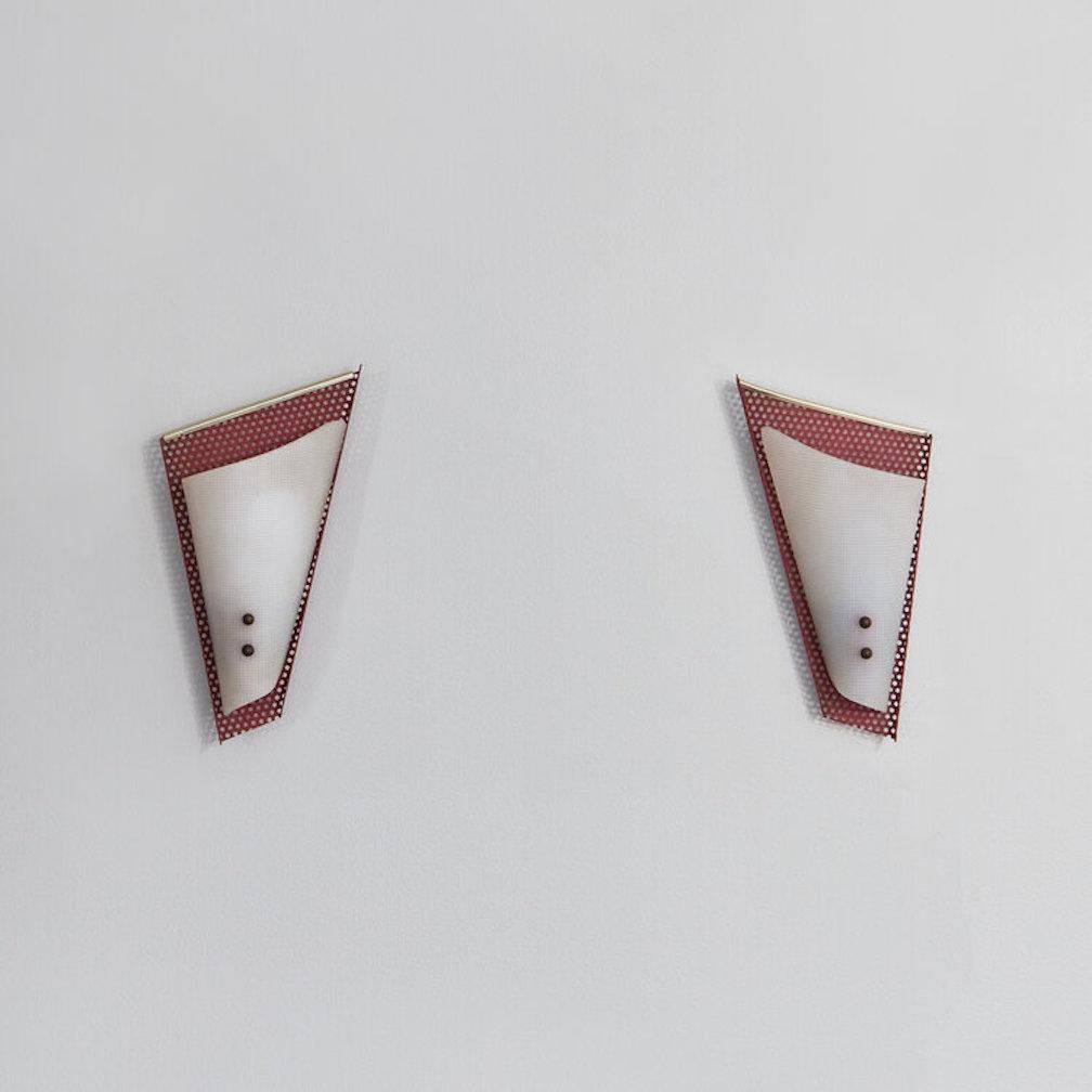 A pair of geometric wall sconces in the style of Jacques Biny. These sconces feature a red perforated metal backing that curves slightly outward to frame a lucite shade attached with brass hardware. When lit, these sconces emanate light in a playful