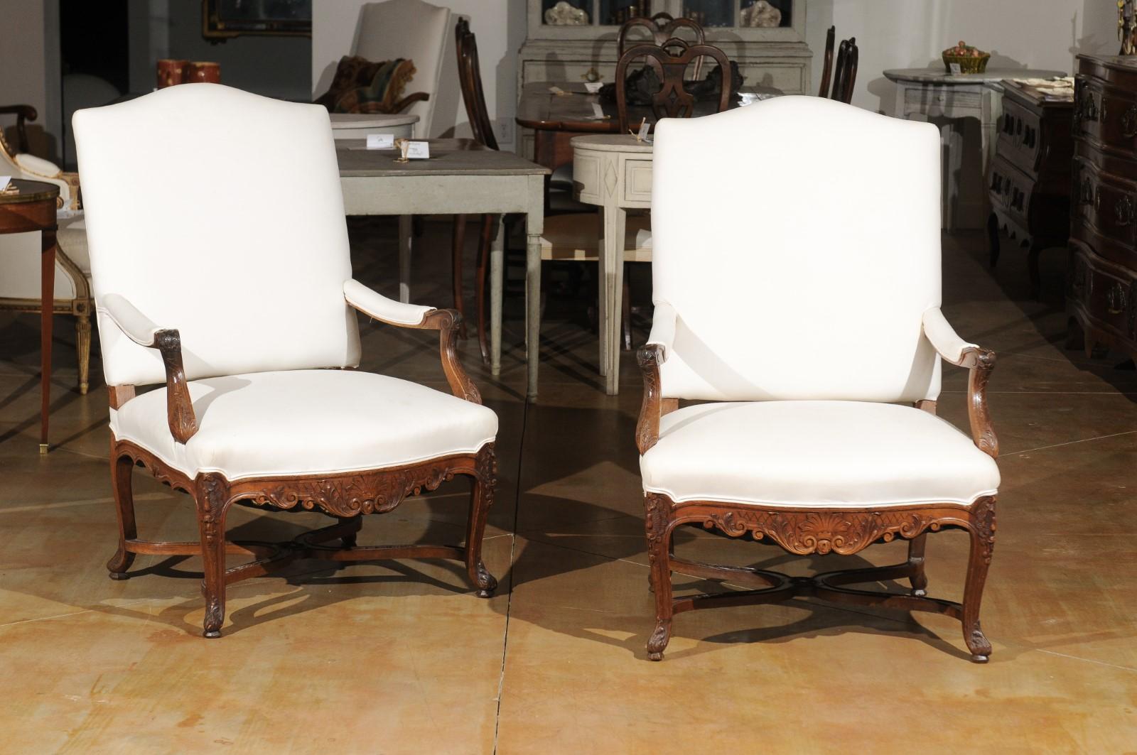 A pair of French walnut Régence style armchairs from the 19th century, with carved foliage motifs, X-form cross stretchers and new upholstery. Created in France during the 19th century, each of this pair of fauteuils features a slanted camelback,