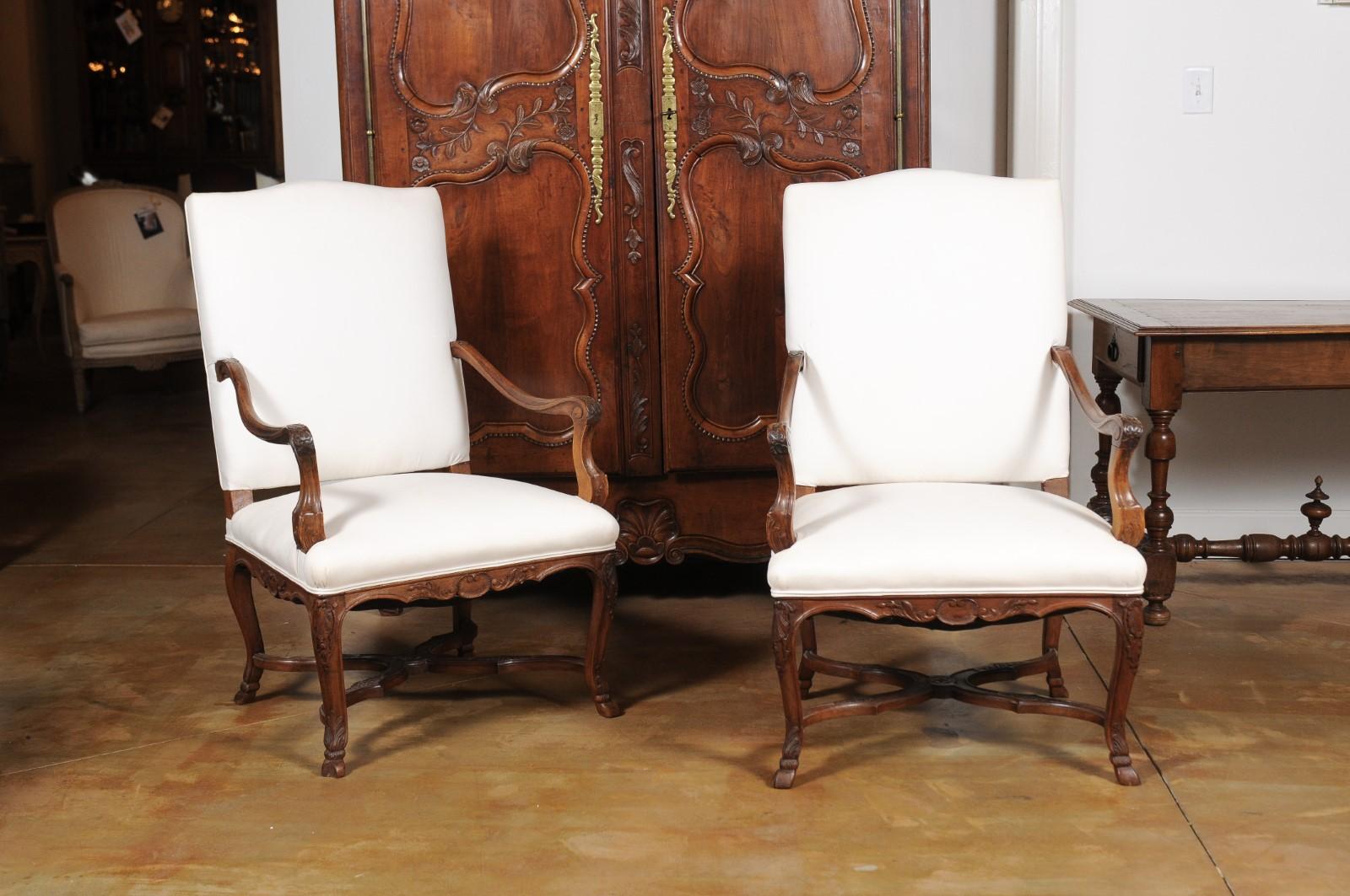 A pair of French Régence style walnut armchairs from the 19th century, with carved foliage, hoofed feet, cross stretcher and new upholstery. Created in France during the 19th century, each of this pair of fauteuils features a slightly slanted back