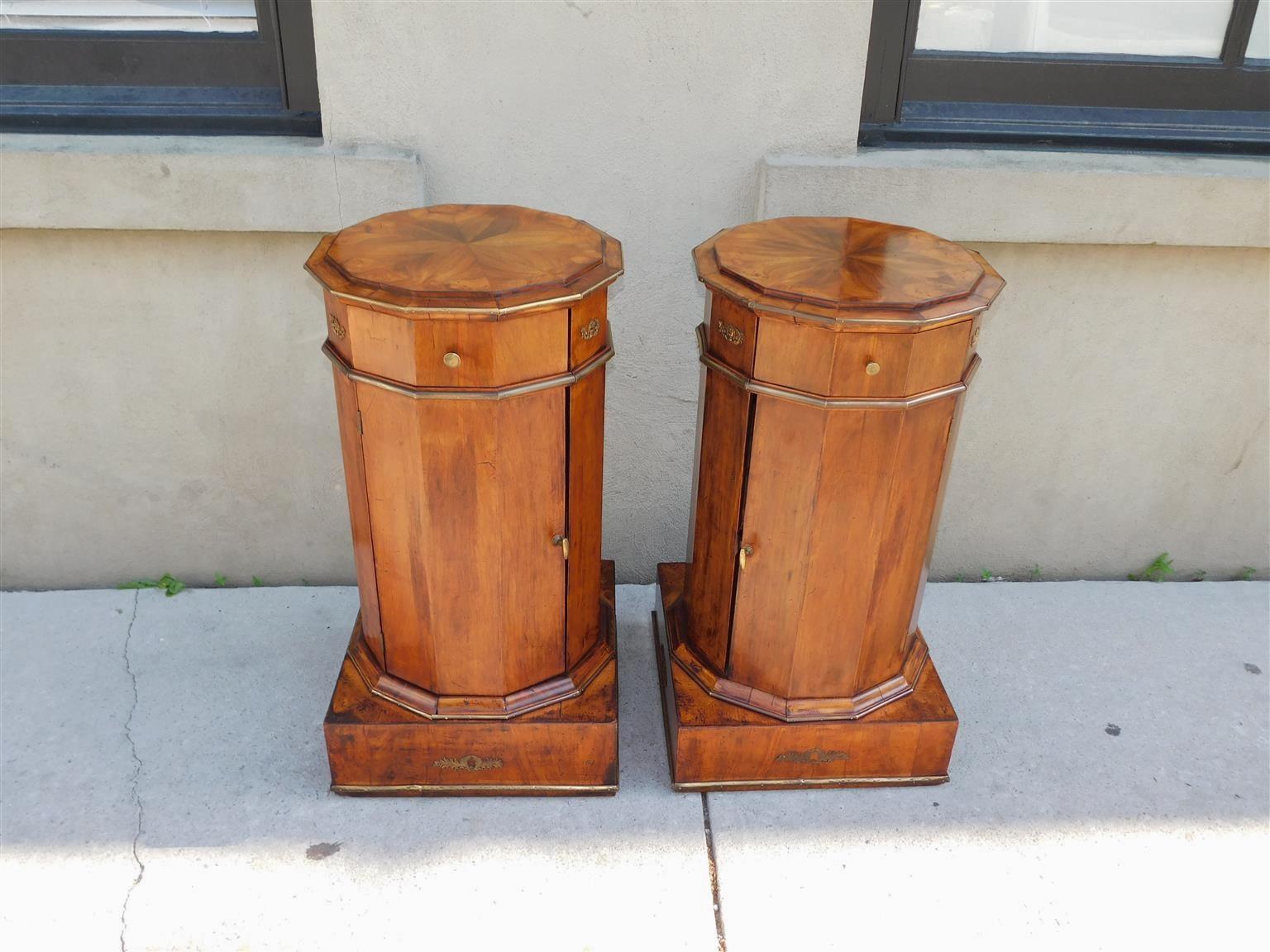 Pair of French Regency book matched mahogany foliage urn ormolu commodes with flanking brass knob single drawers, brass handle cabinets with interior shelf, and resting on squared brass trimmed bases.  Early 19th Century 