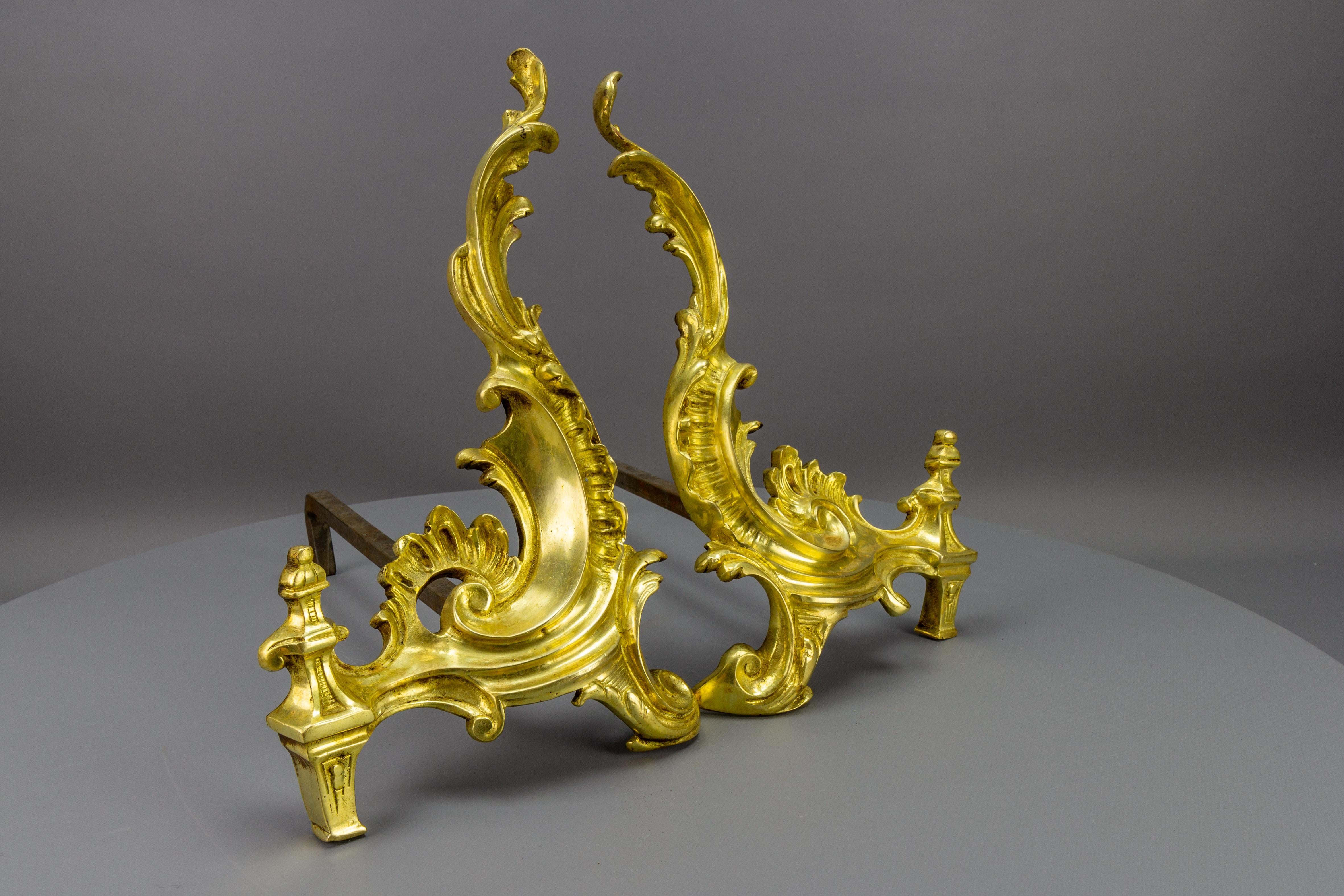 A pair of antique Louis XV or Rococo style firedogs made of gilt bronze and iron, France, circa the 1910s. 
These marvelous firedogs or andirons feature gilt bronze construction of typical Rococo design, of scroll shape with scrolling foliate and