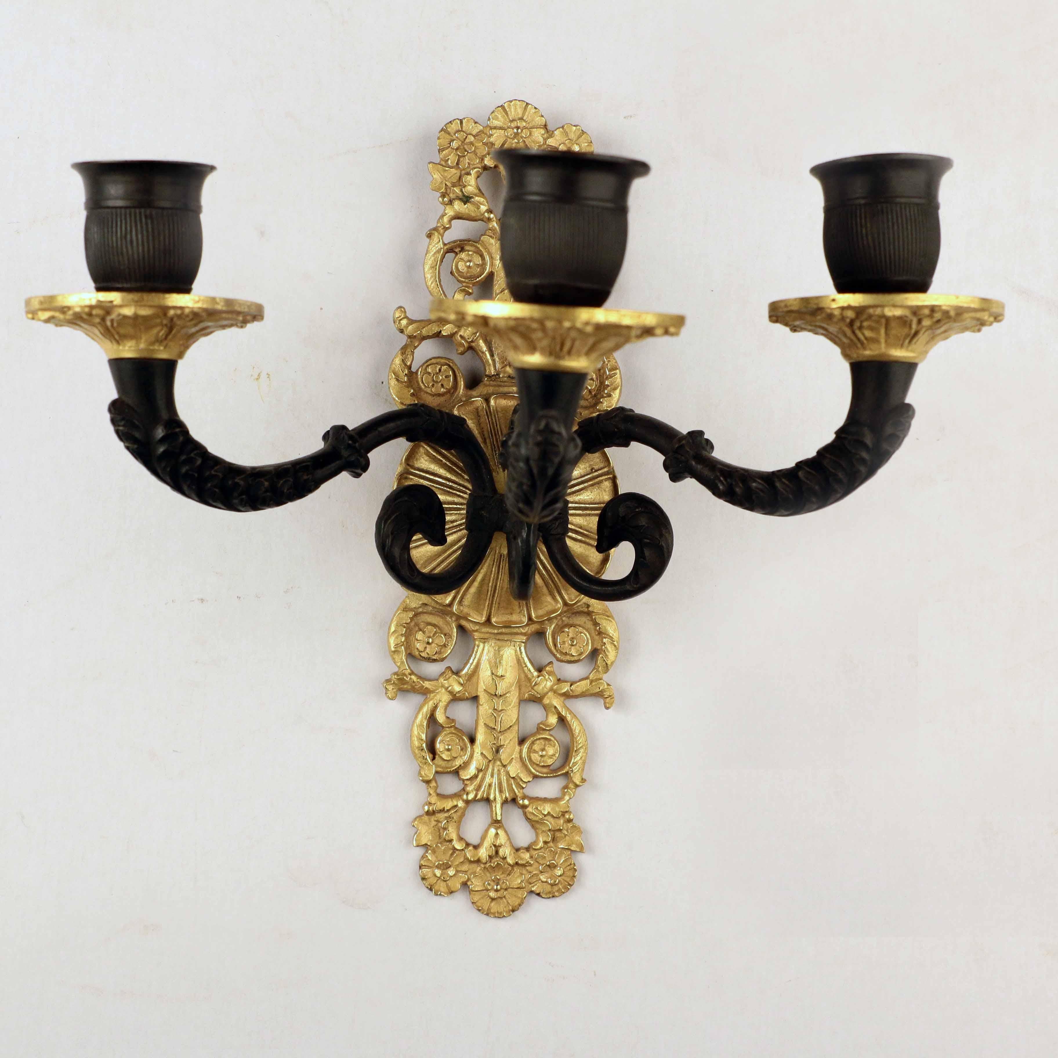 These good quality sconces in mat black and gilt bronze are pierced with scrolls and flower heads and with drip pans and arms cast with stylized acanthus. The two-tone gilding and handmade nut suggest a date no later than the Belle Époque. This