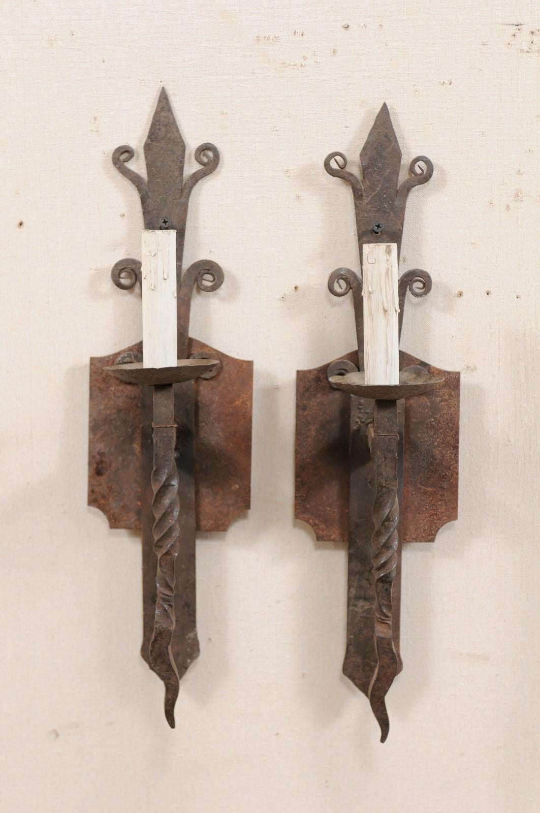 A pair of French single-light iron sconces from the mid-20th century. This vintage pair of French sconces each feature a vertically positioned and stylized back plate with curled iron accents and speared top. A single, straight arm projects from the