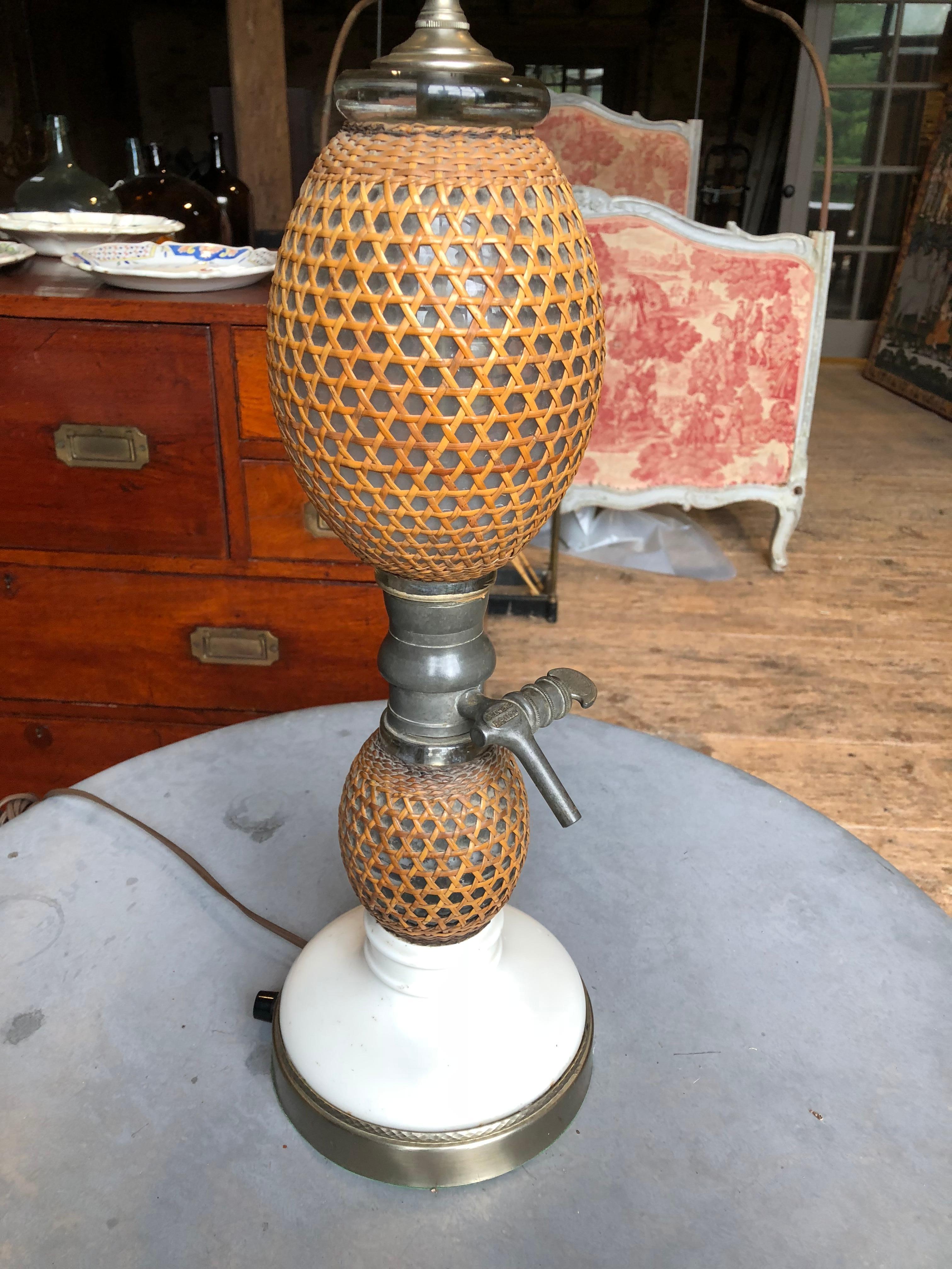 A charming pair of French lamps made from 19th century bistro seltzer siphons, with wicker covered glass reservoirs and pewter spigots on a white porcelain base. The lamps are currently mounted on nickel plated bases with a surface mounted switch on