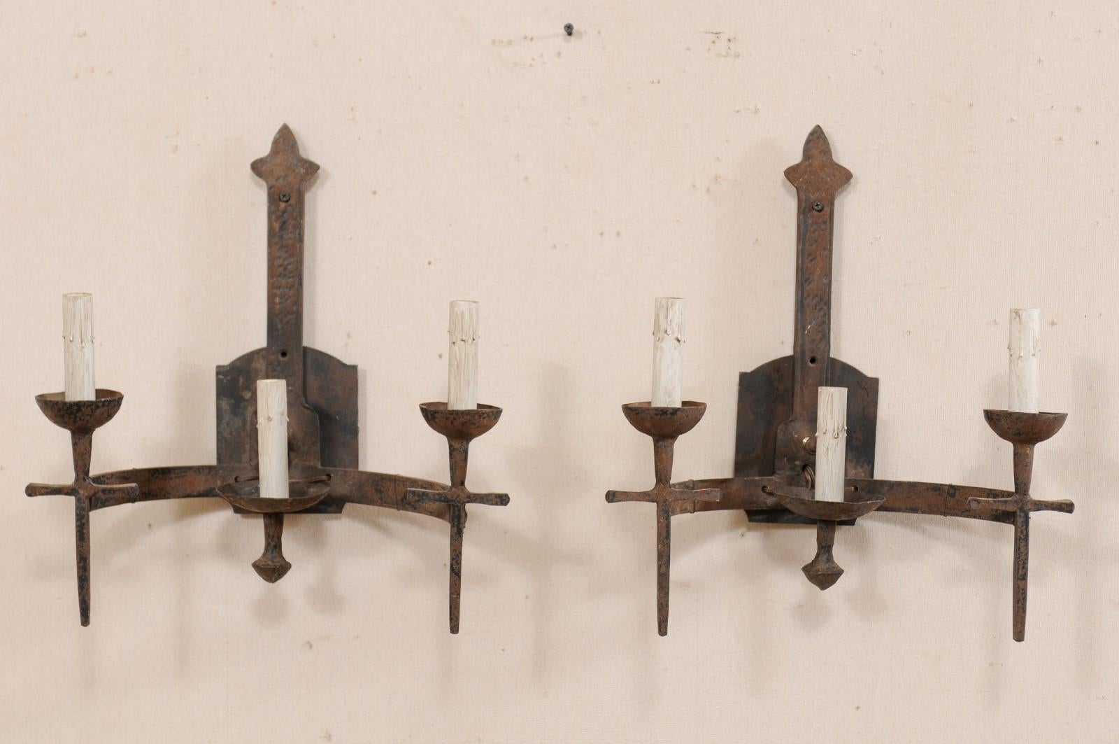 A pair of French three-light iron sconces from the mid-20th century. These vintage French hand forged sconces each have two torch-style lights set at the farthest ends of a horizontally set c-shaped arm, flanking a shorter profiled central light