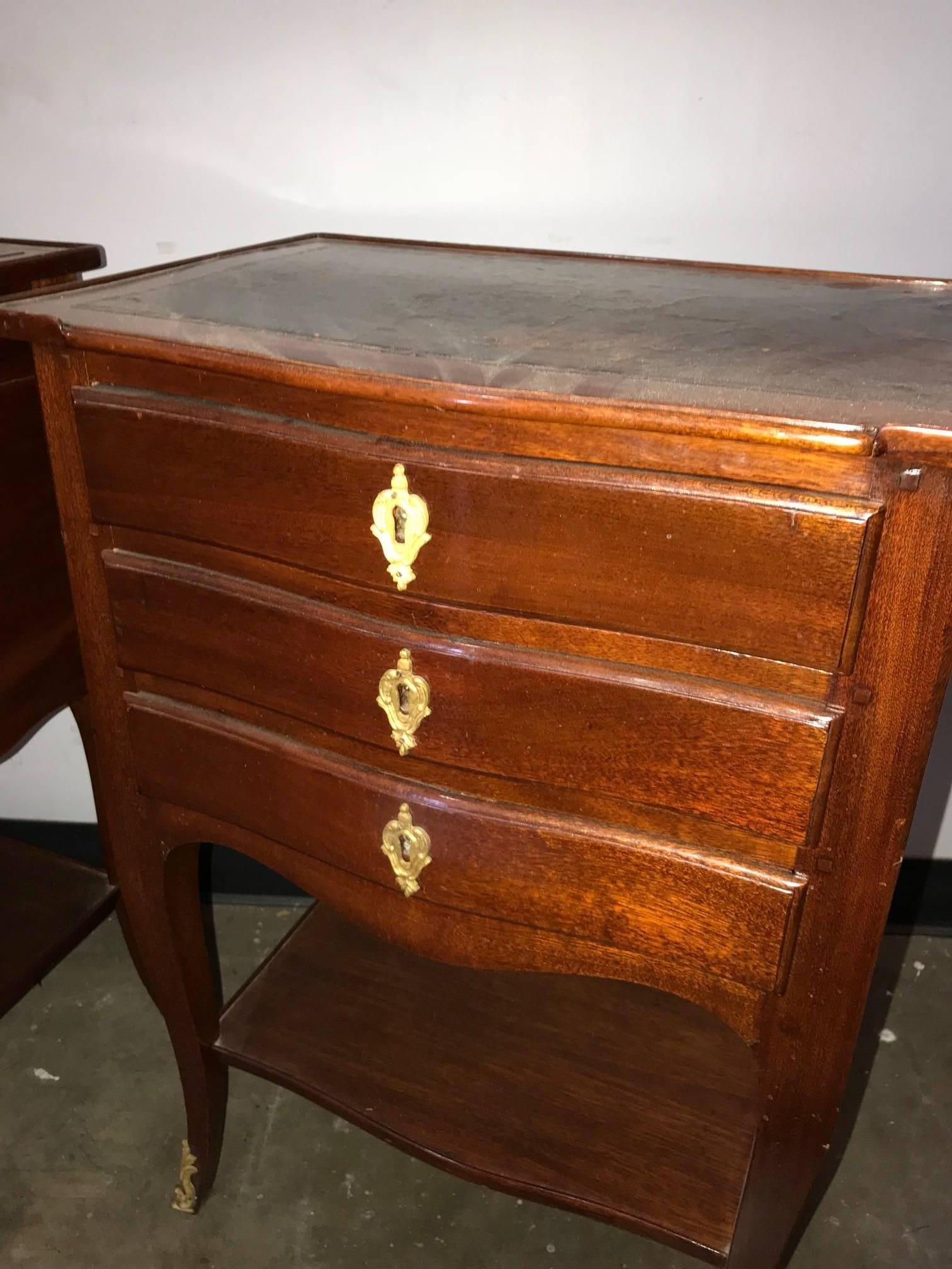 Attractive pair of French Louis XV style mahogany shaped front side tables or night stands fitted with three drawers with gold gilt ormolu escutcheons and having a lower tier. The entire on slender cabriole legs ending in gilded bronze