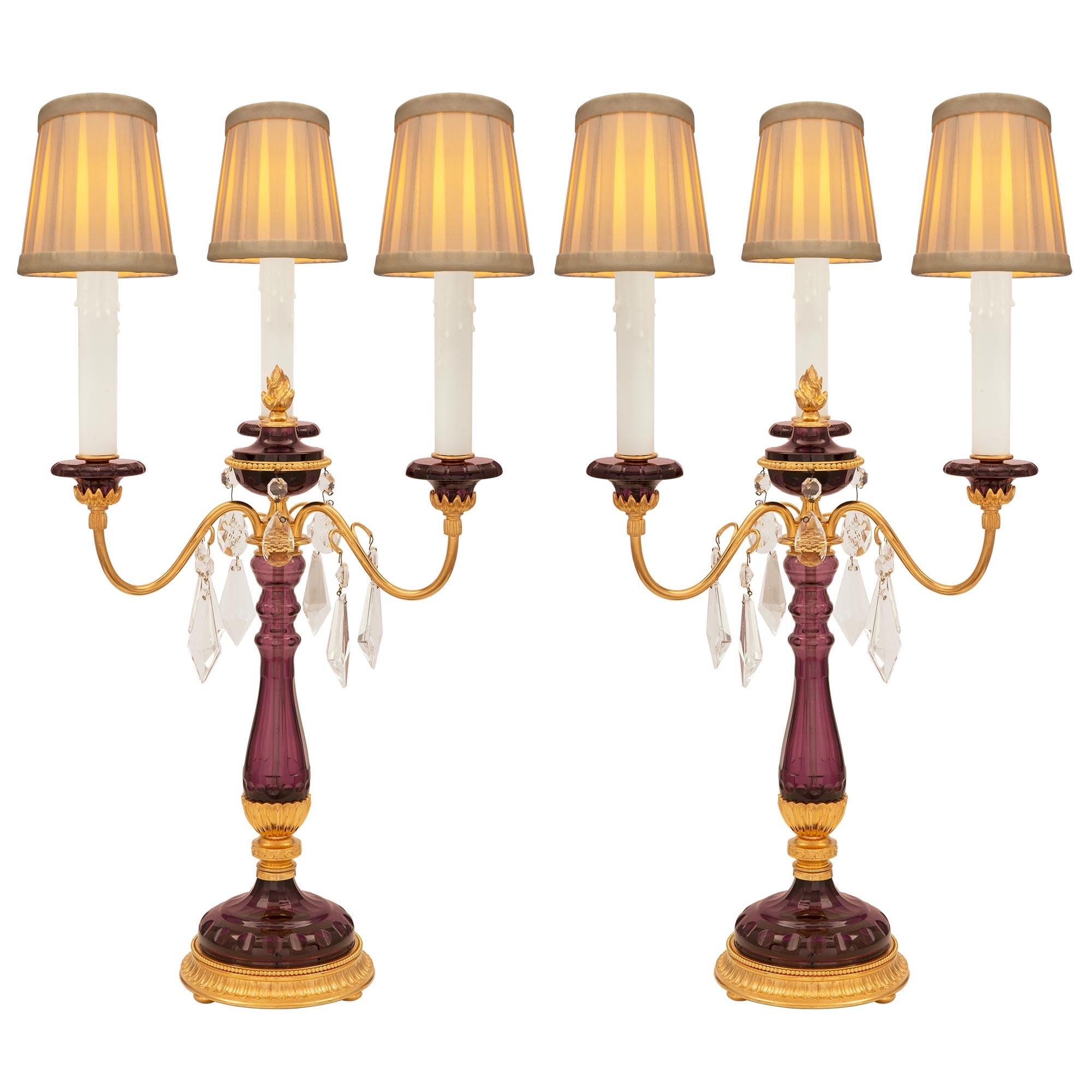 A beautiful and unique pair of French turn of the century Louis XVI st. amethyst colored glass, ormolu, and crystal candelabra lamps. Each three arm lamp is raised by an elegant circular ormolu base with bun feet and a fine wrap around curved reeded