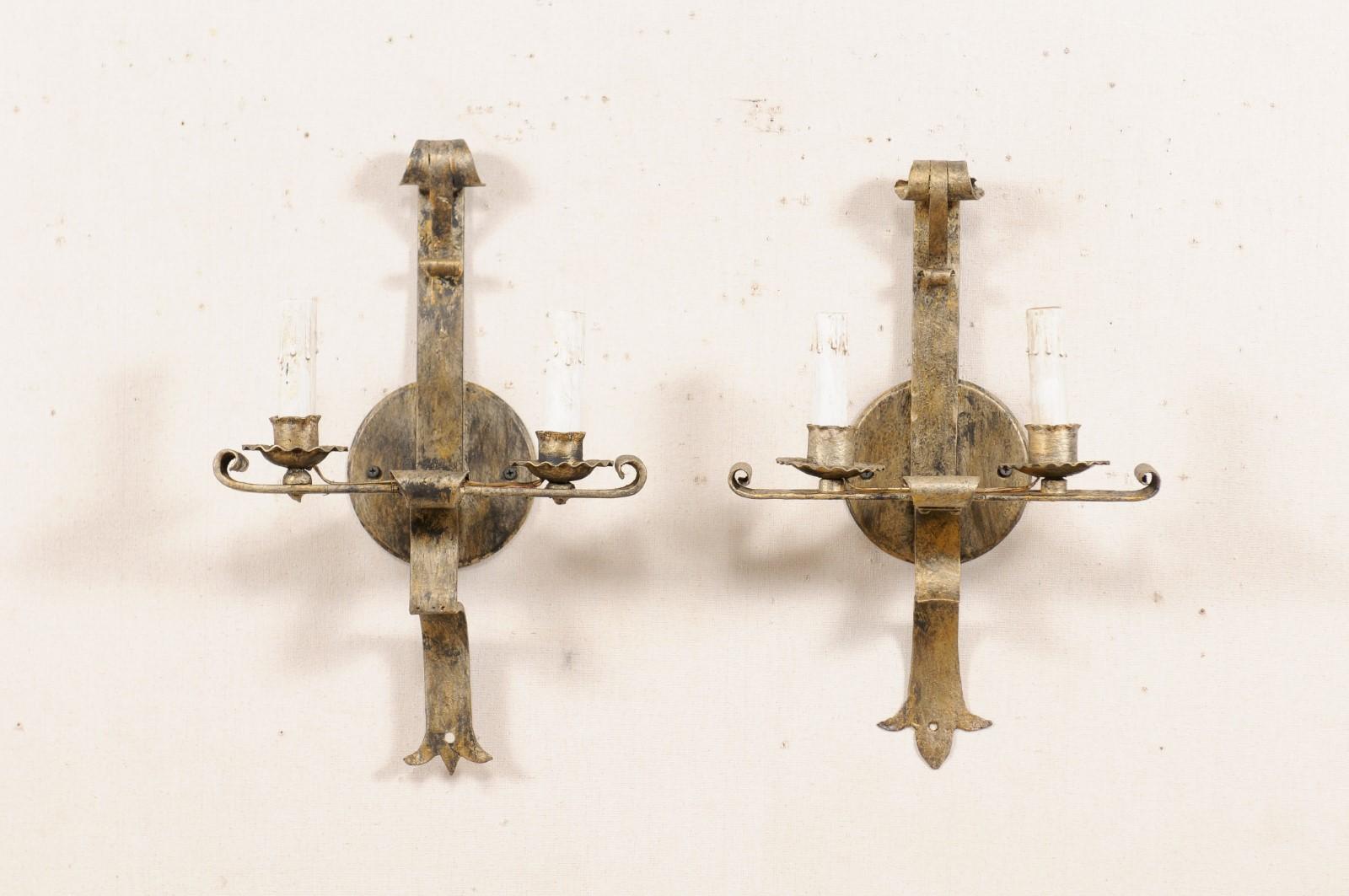 A pair of French two-light gold-tone iron sconces from the mid-20th century. These vintage hand-forged sconces from France each feature a horizontally placed bar, with up-turned tips at each end, providing support to the two ruffled iron bobèches