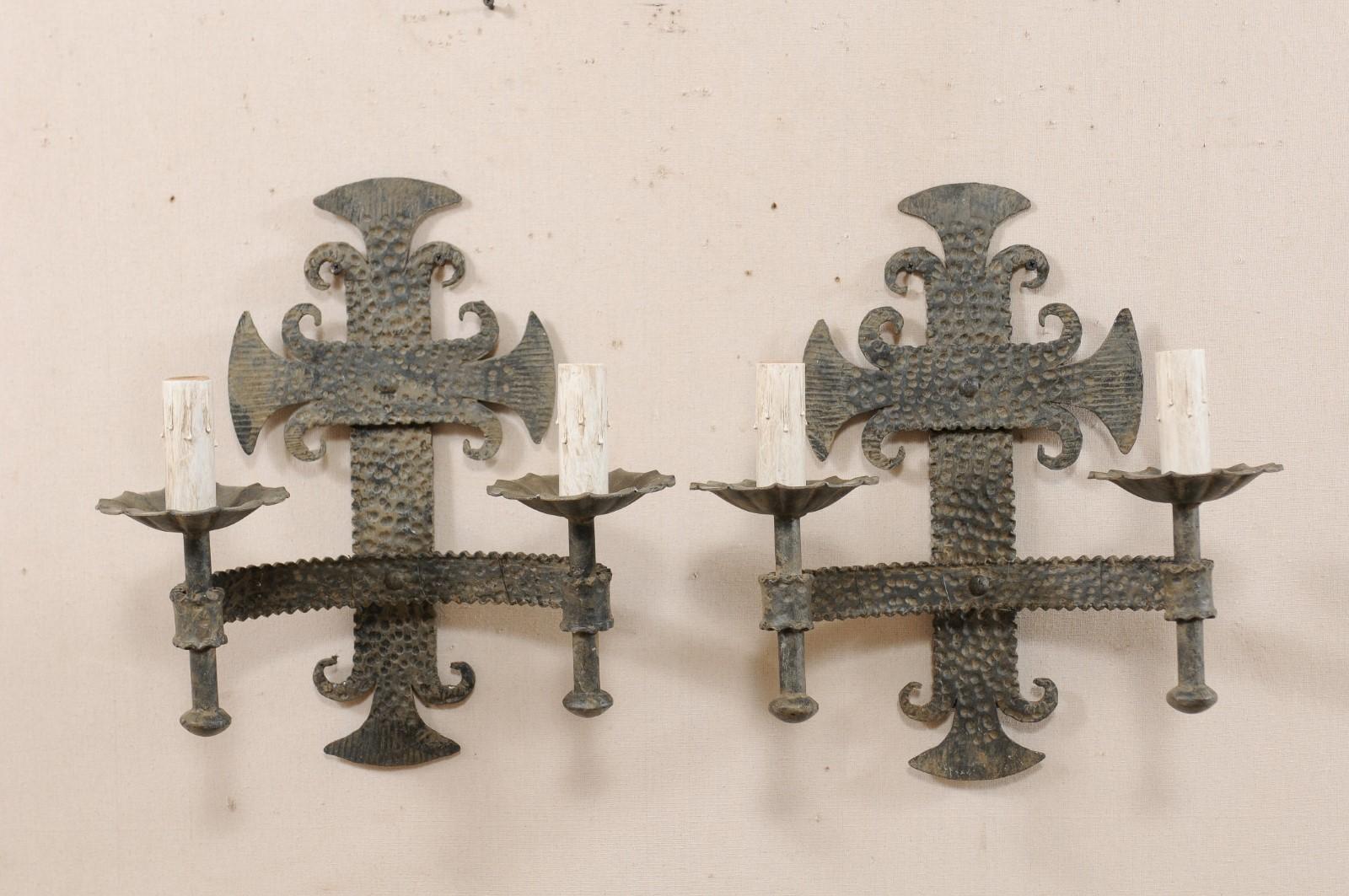 Pair of French two-light iron sconces from the mid-20th century. This vintage pair of sconces from France each feature a stylized fleur de Lis back-plate with hammered texturing, from which a c-shaped arm extends out supporting the torch style