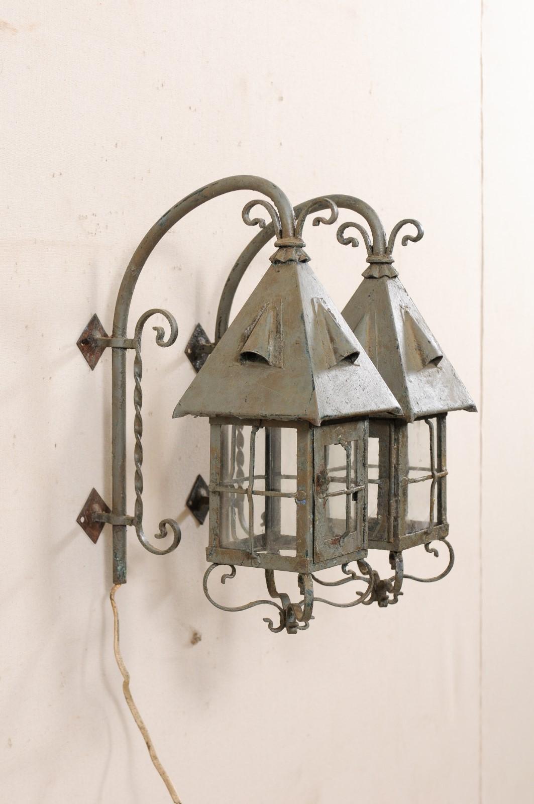 A pair of French painted iron wall-mounted lanterns from the mid-20th century. This pair of vintage sconces from France each have lantern style bodies which are extended outward from the wall by a curved, overhanging back post, each decorated with