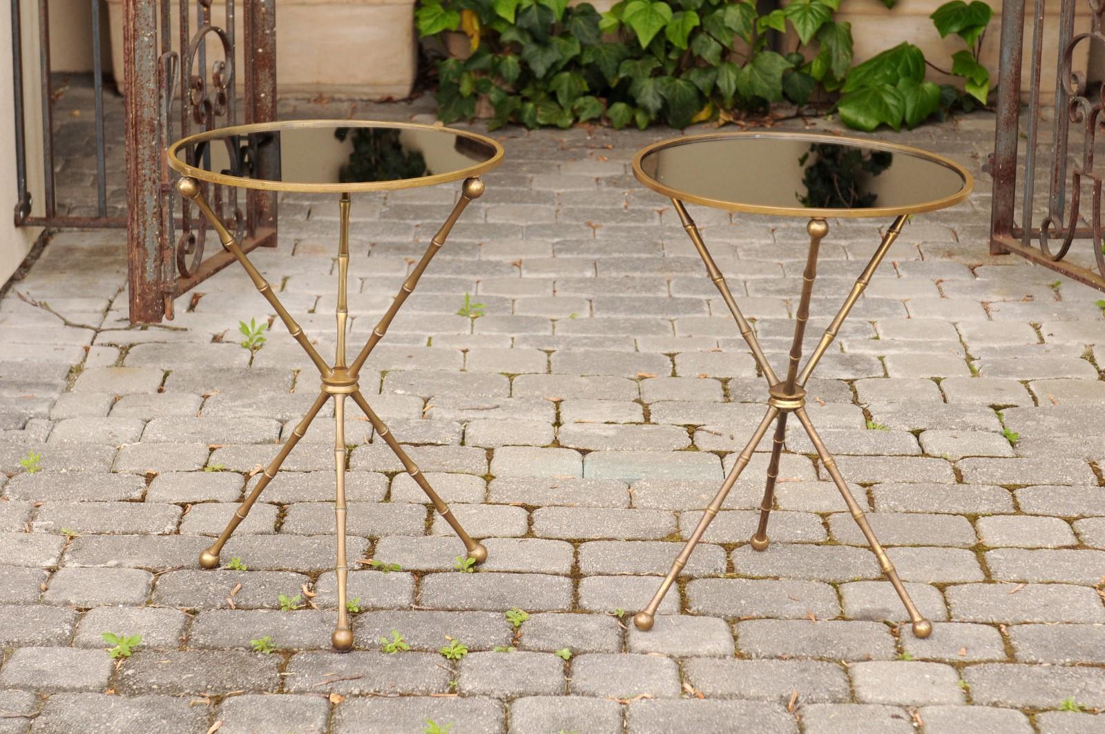 A pair of vintage French Maison Jansen style round side tables with faux-bamboo brass base and black mirrored tops from the midcentury. Born in France during the mid-century period, each of this pair of side tables was produced in the manner of