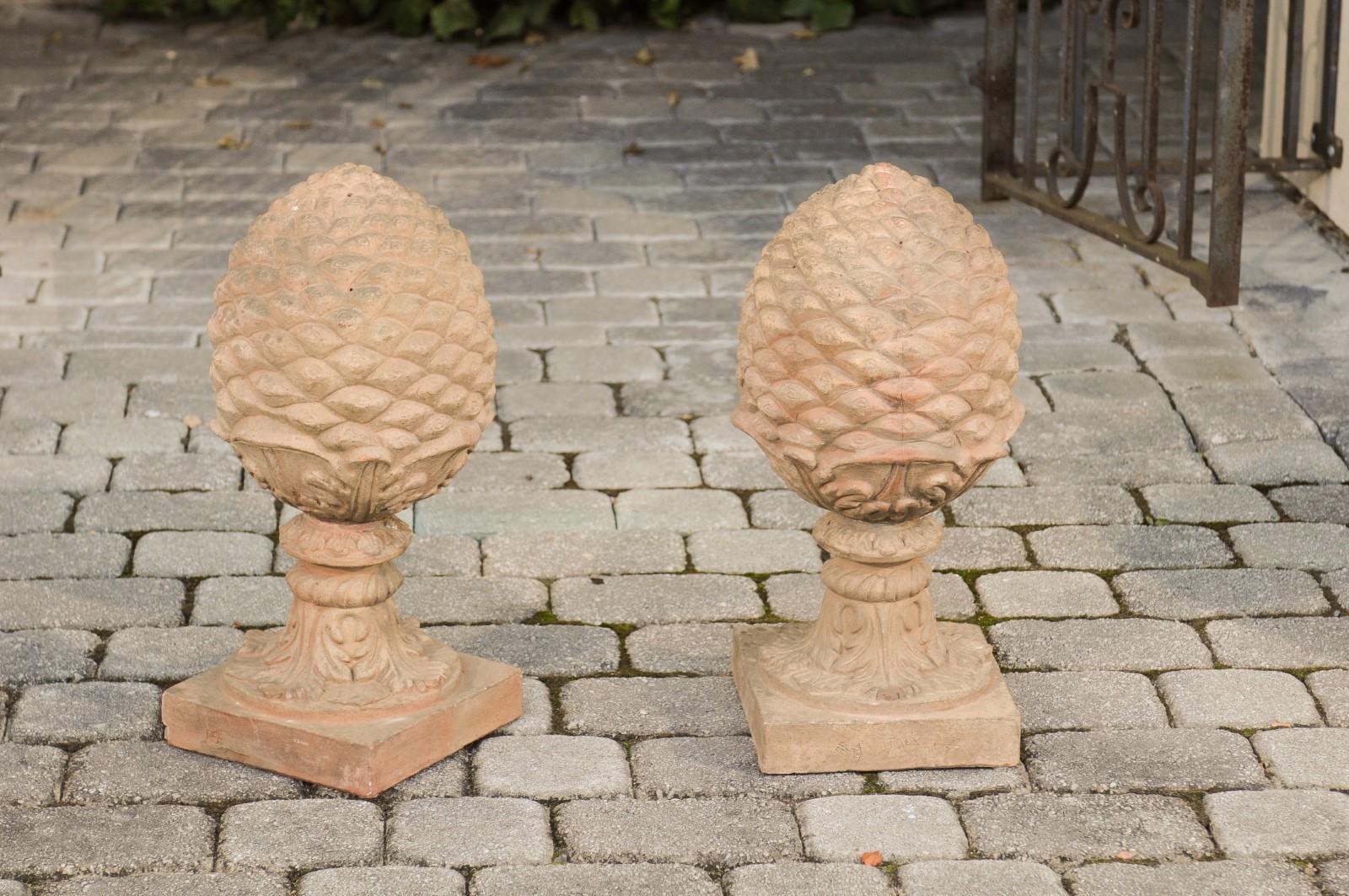 A pair of French vintage painted terracotta pinecone finials with acanthus leaves and square bases. Born in France during the first half of the 20th century, each of this pair of terracotta finials depicts pinecones supported by acanthus leaves