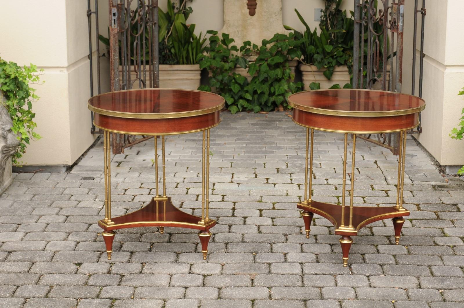 A pair of French mahogany and brass circular tables from the mid-20th century, with faux bamboo supports, tripartite shelves and toupie feet. Born in France during the mid-century period, each of these two exquisite tables features a circular