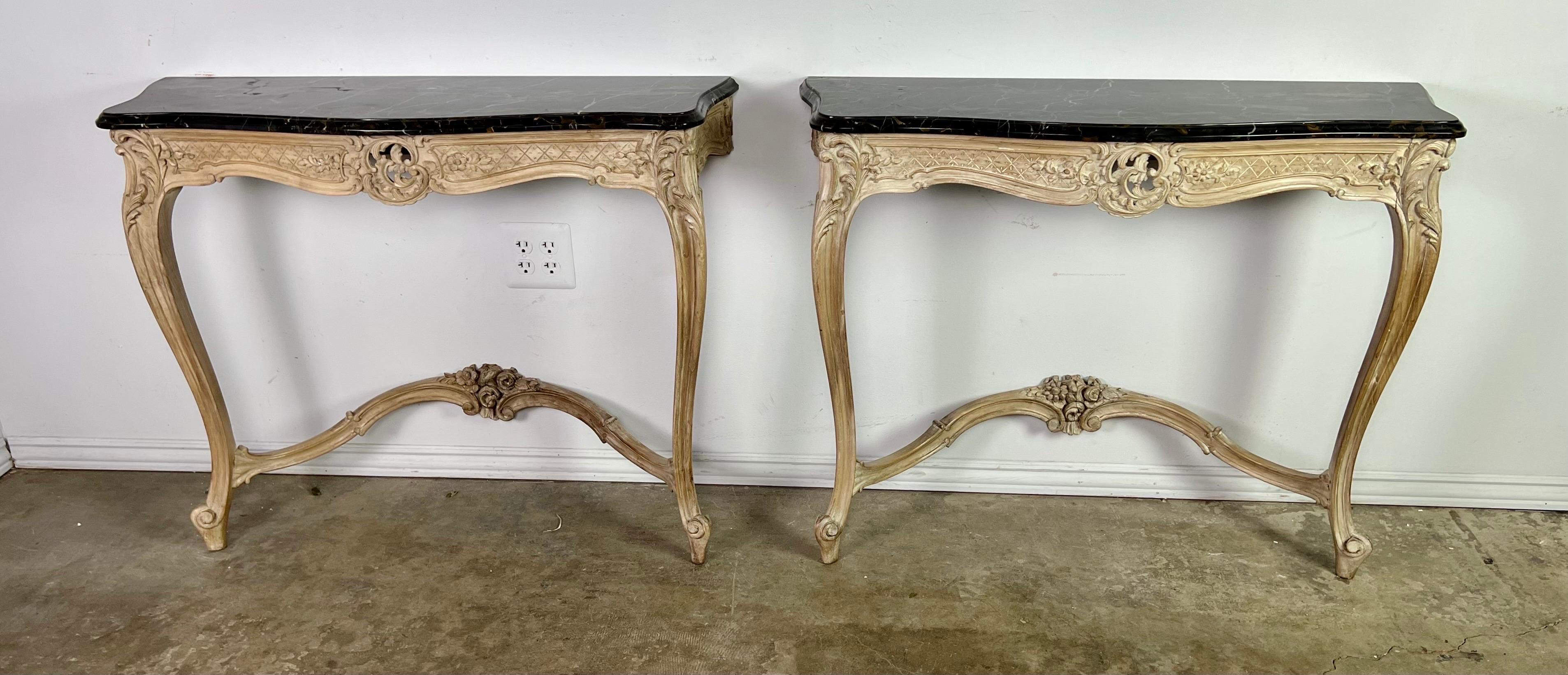 Pair of French Louis XV style carved wood consoles with original black marble tops. There is carved roses at the bases of the consoles that each stand on two cabriole legs that end in rams head feet. There is more carving at the top center of the
