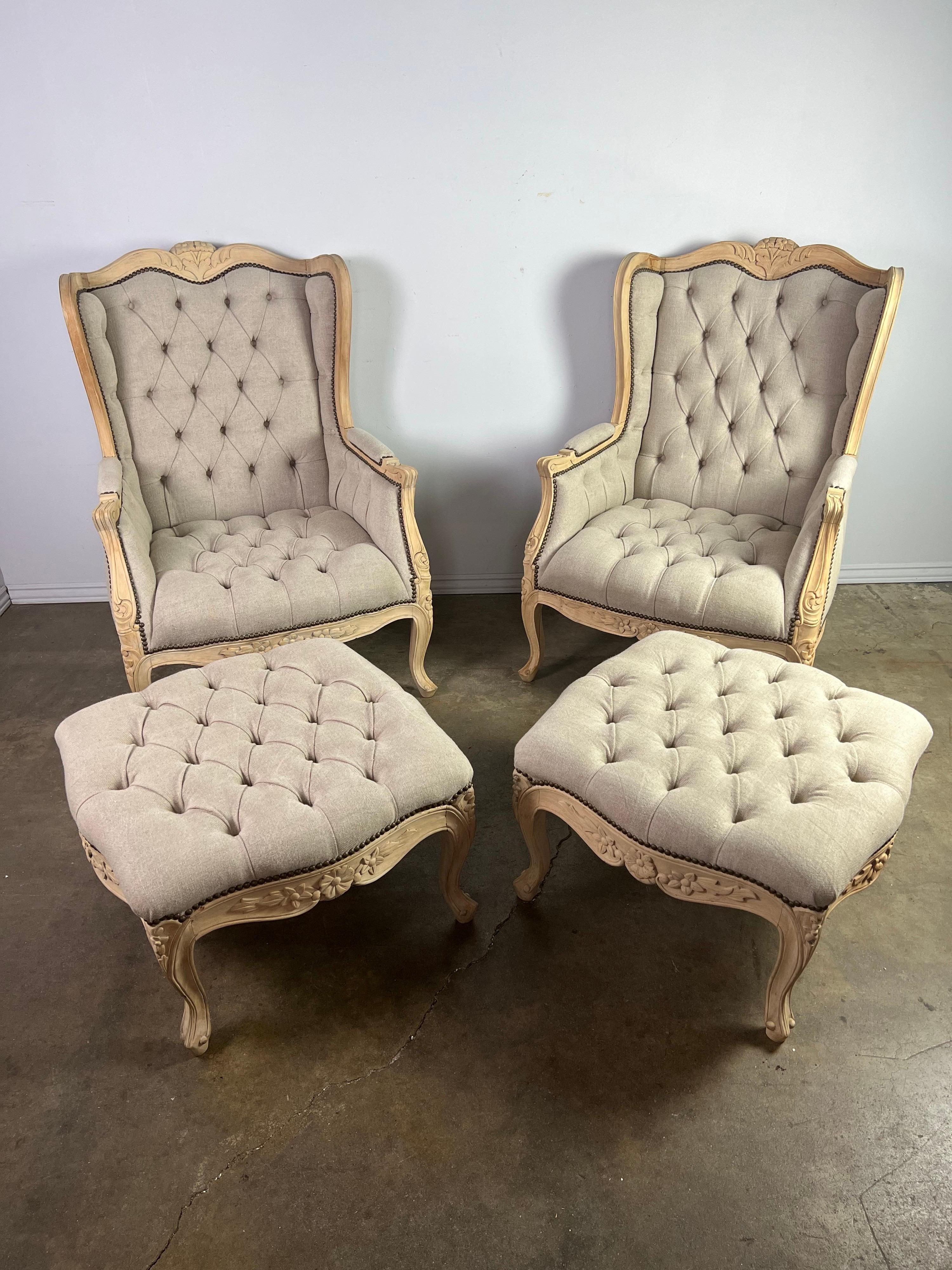 Pair of French carved bleached walnut wingback armchairs w/ matching ottomans. The chairs and ottomans stand on cabriole legs with rams head feet. The chairs are newly upholstered in a washed tufted Belgium linen with nailhead trim detail.

The