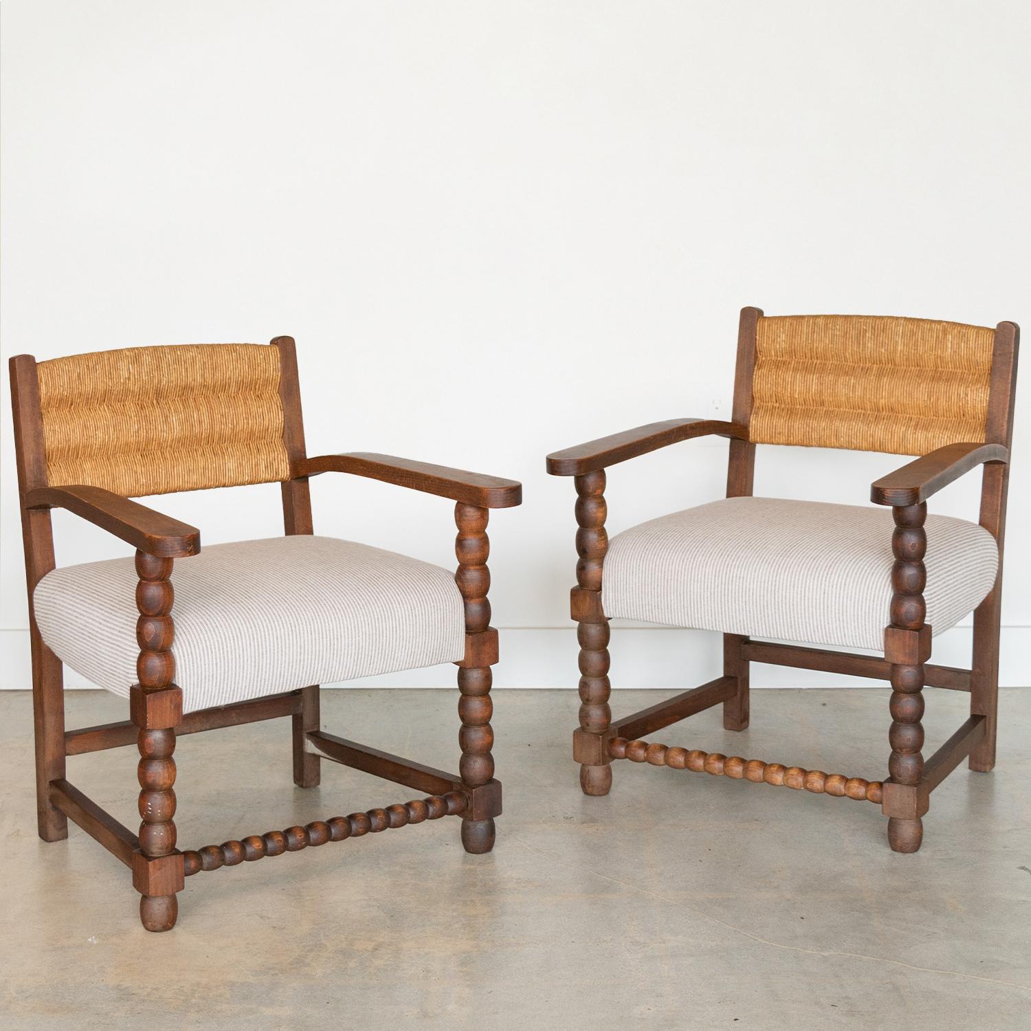 Wonderful pair of carved wood armchairs from France, 1940s. Beautiful carved bobbin wood legs and frame with dark stain. Original woven rush backs. Newly upholstered seat in pale striped cream and brown linen blend.