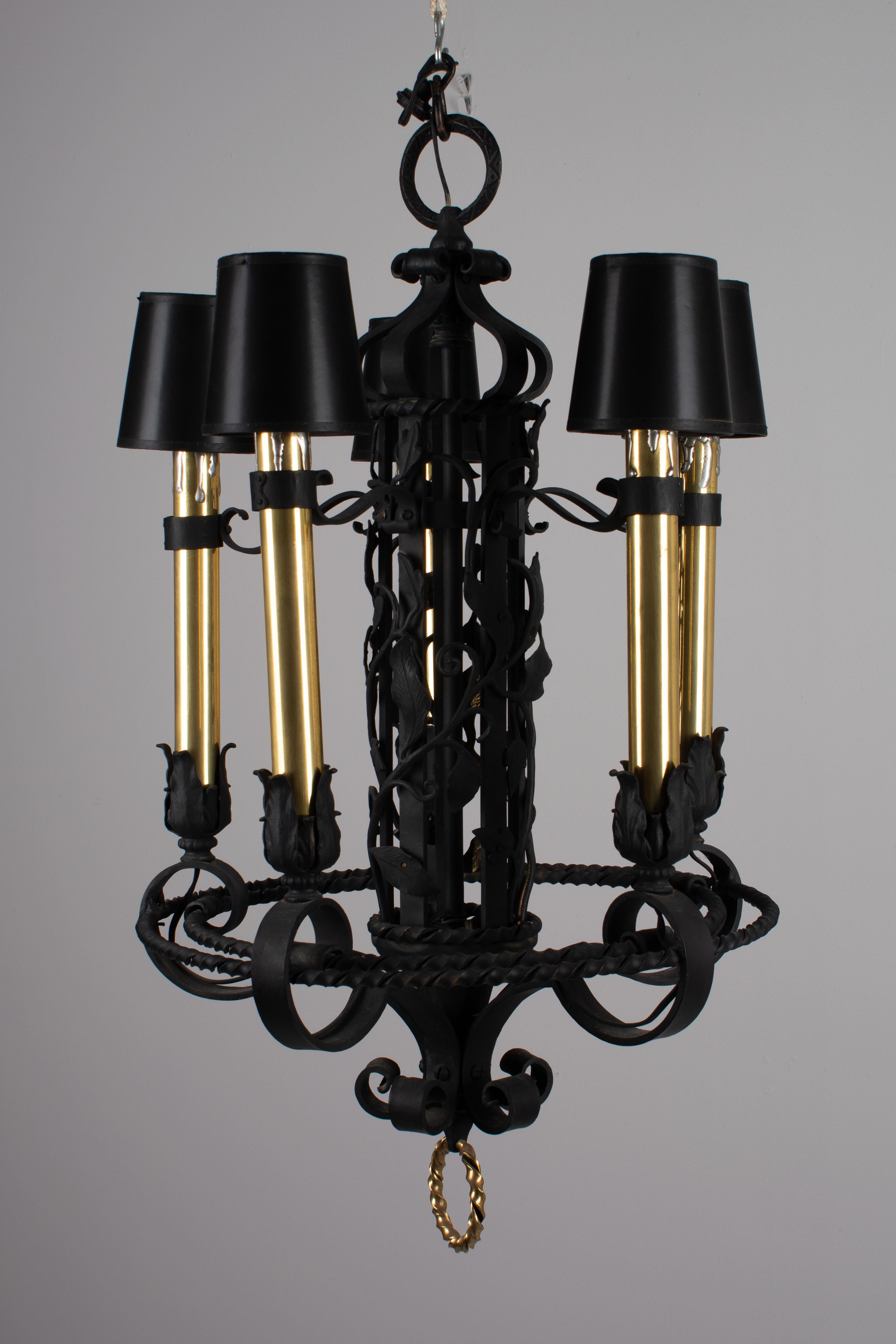 A pair of large French five-light wrought iron chandeliers. Beautifully crafted iron work with a central column entwined in vines and leaves and surrounded by five tall  solid brass candles. Old black shades with gold foil lining might need to be