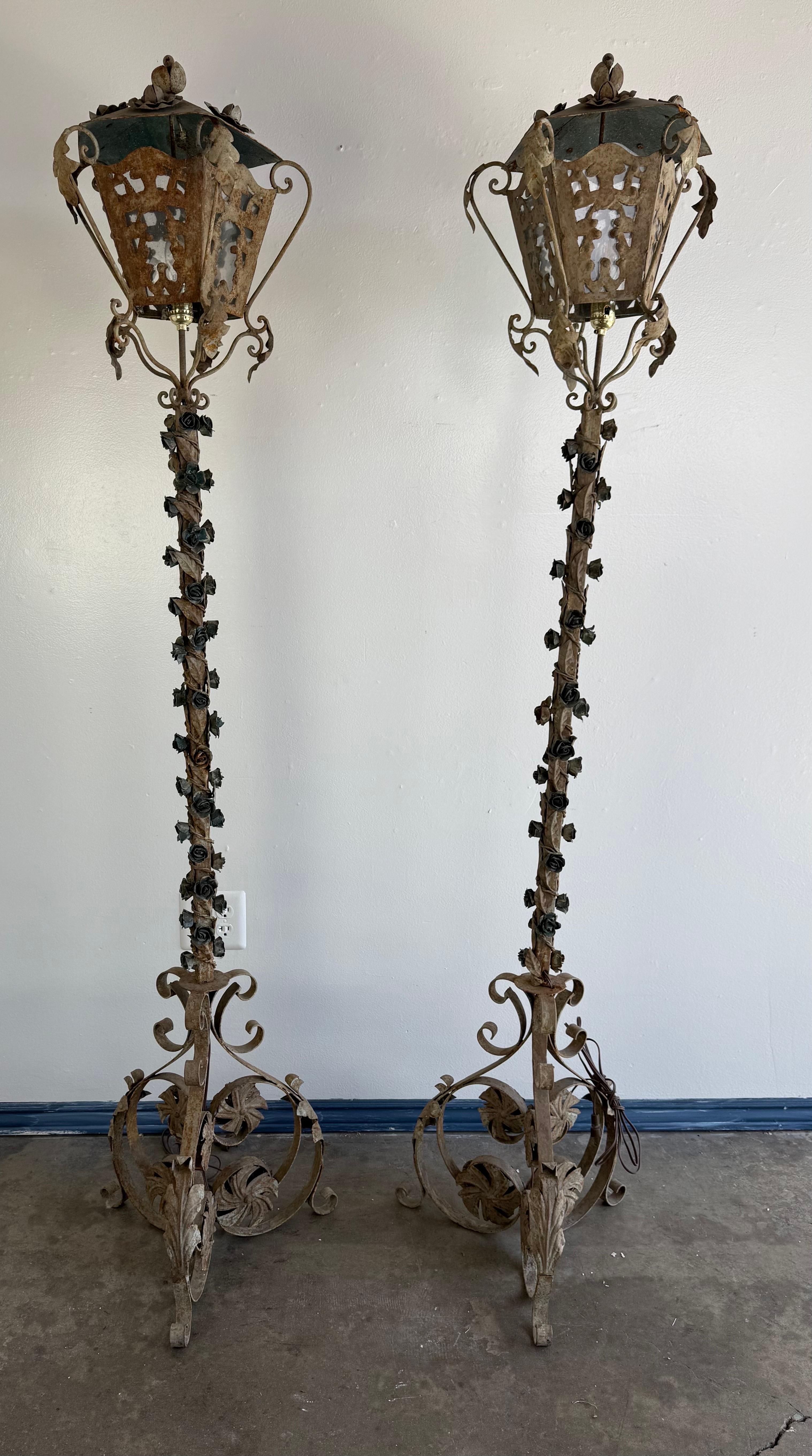 Il s'agit d'une paire de lanternes de sol vintage en fer forgé à l'esthétique ornée et rustique. Le motif présente des volutes complexes et des motifs floraux, y compris des accents de rose grimpant le long des tiges élancées. Les lanternes ont une