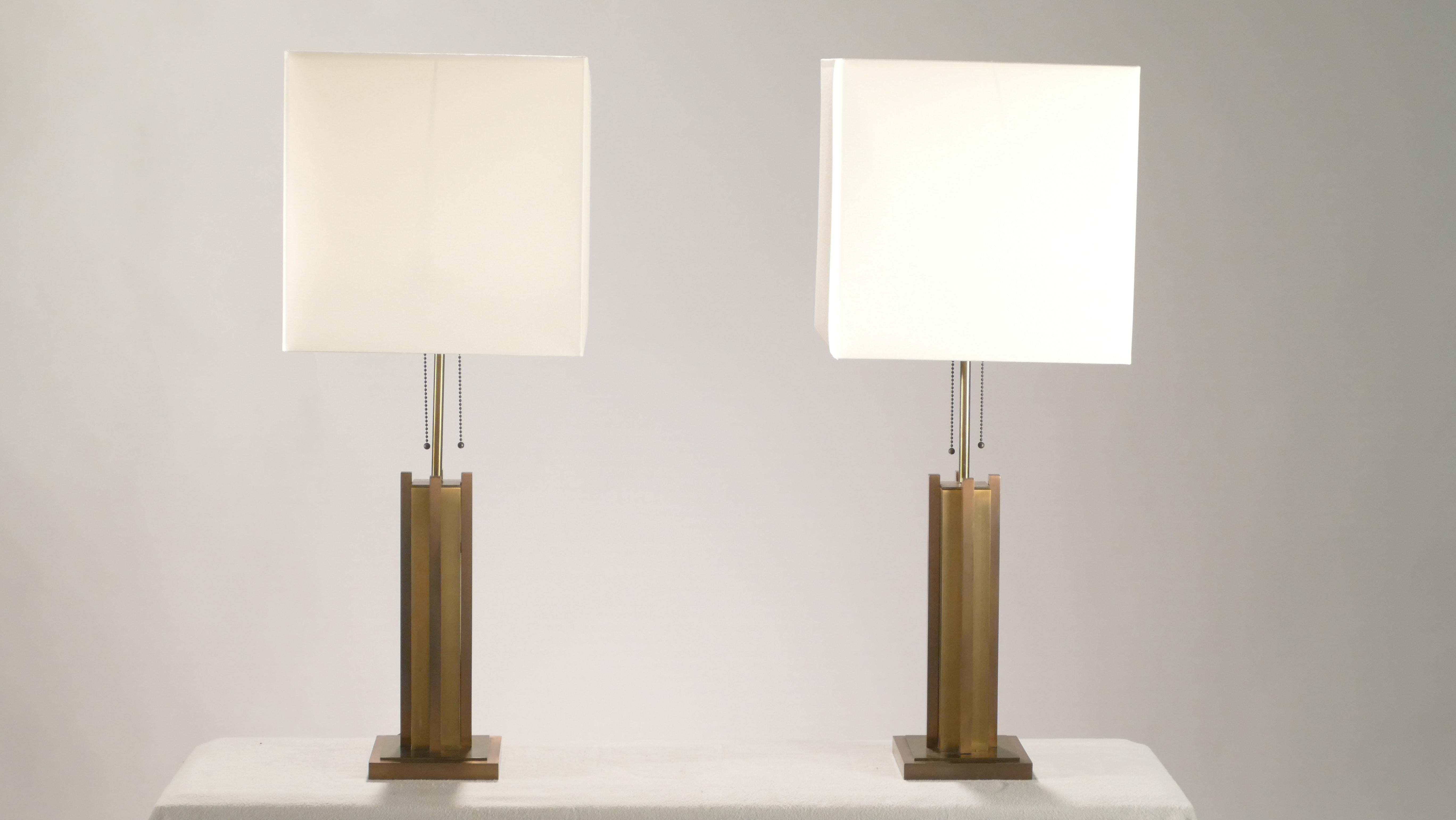 This pair of brass and bronze lamps features a beautiful metal combination of brass and bronze, their complementary shades enhanced by the base’s stunning architectural design. Topped with creamy white lampshades, the lamps have a professional and