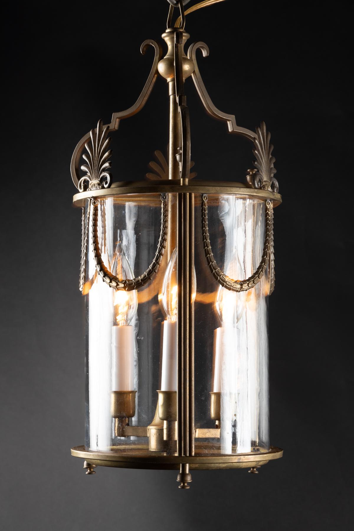 These small cylindrical lanterns are decorated with streams of garlands hanging down from the upper metal rim of the lantern. A trio of palmetto fronds sits atop the metal rim, spaced between three arms that hold the stem of the lantern. The glass