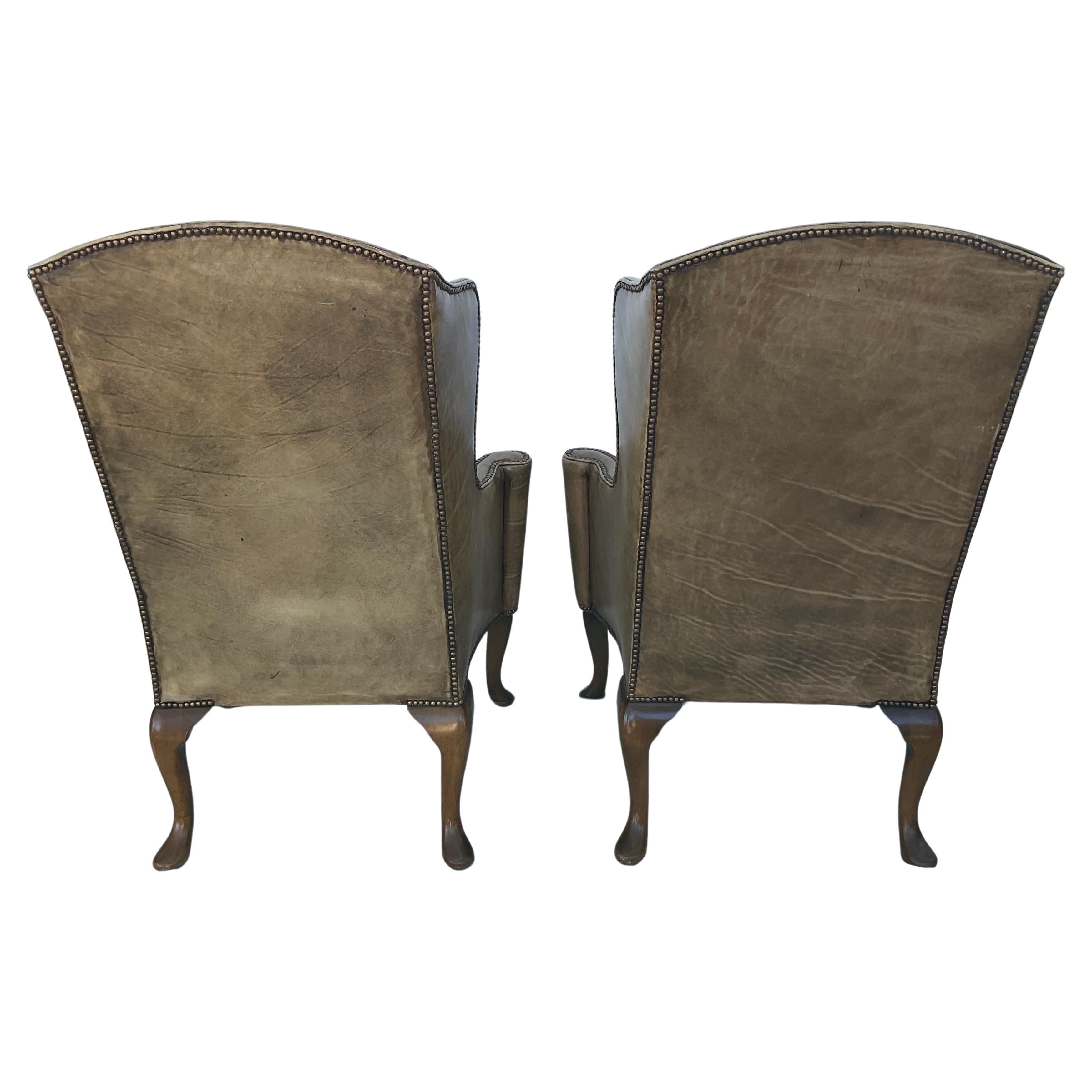 A pair of George I style leather winged armchairs. Each chair features an arched backrest with shaped wings and scrolled arm rests centering a loose cushioned seat, raised on cabriole legs. 