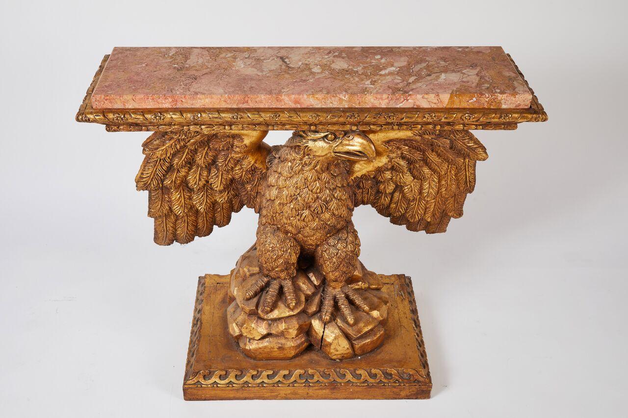 A pair of George II style Kentian carved giltwood eagle consoles from the early 20th century. The rouge marble tops sit within an intricately carved edge border of ribbon and rosettes. The eagles sit proudly upon rocky outcrops. Bold Vitruvian