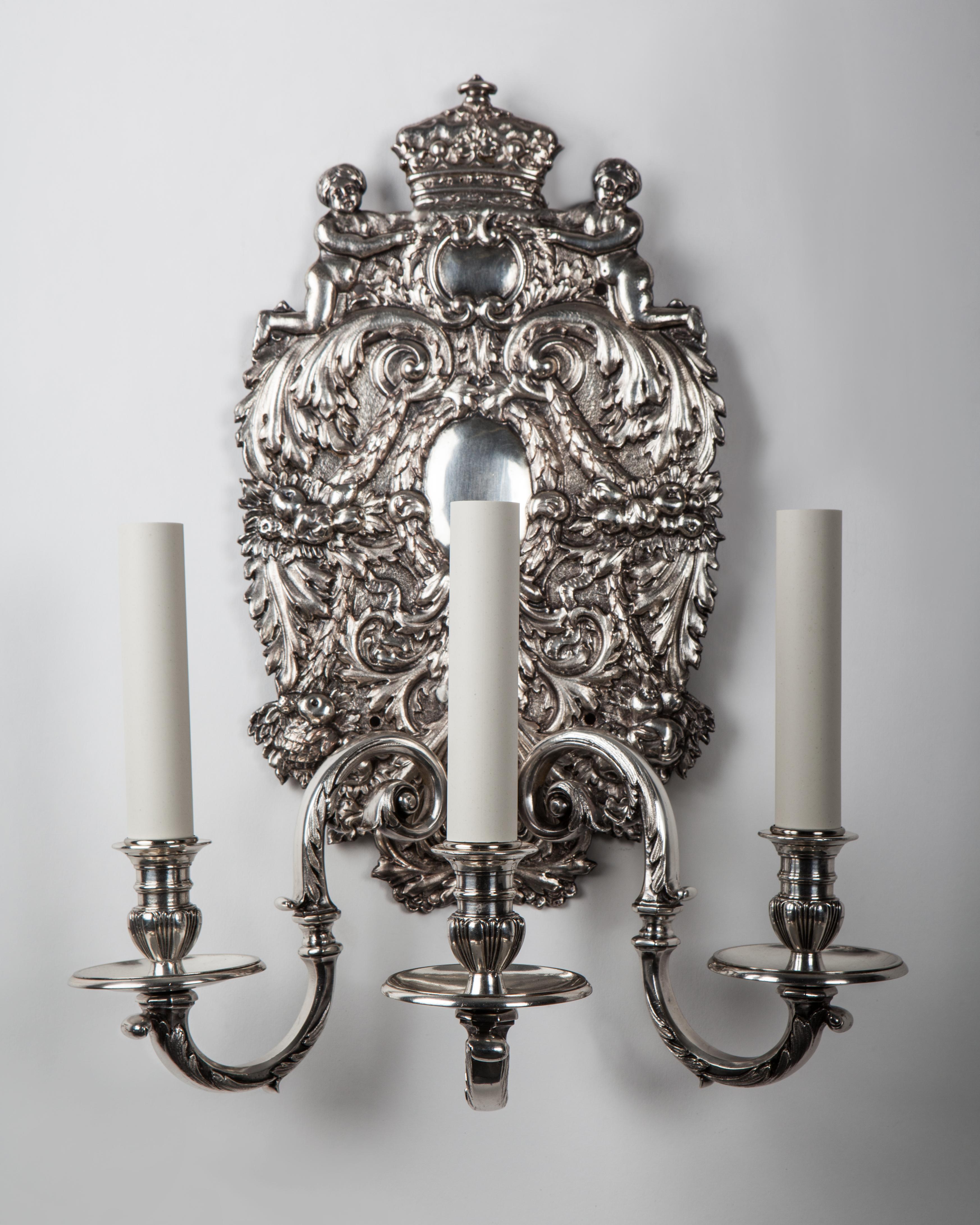 AIS2948
A pair of antique three arm sconces in silverplate over bronze with George II style bas-relief backplates having cherubs holding crowns and foliate details. The S-curving arms supporting gadrooned waxpans and candle cups. Circa