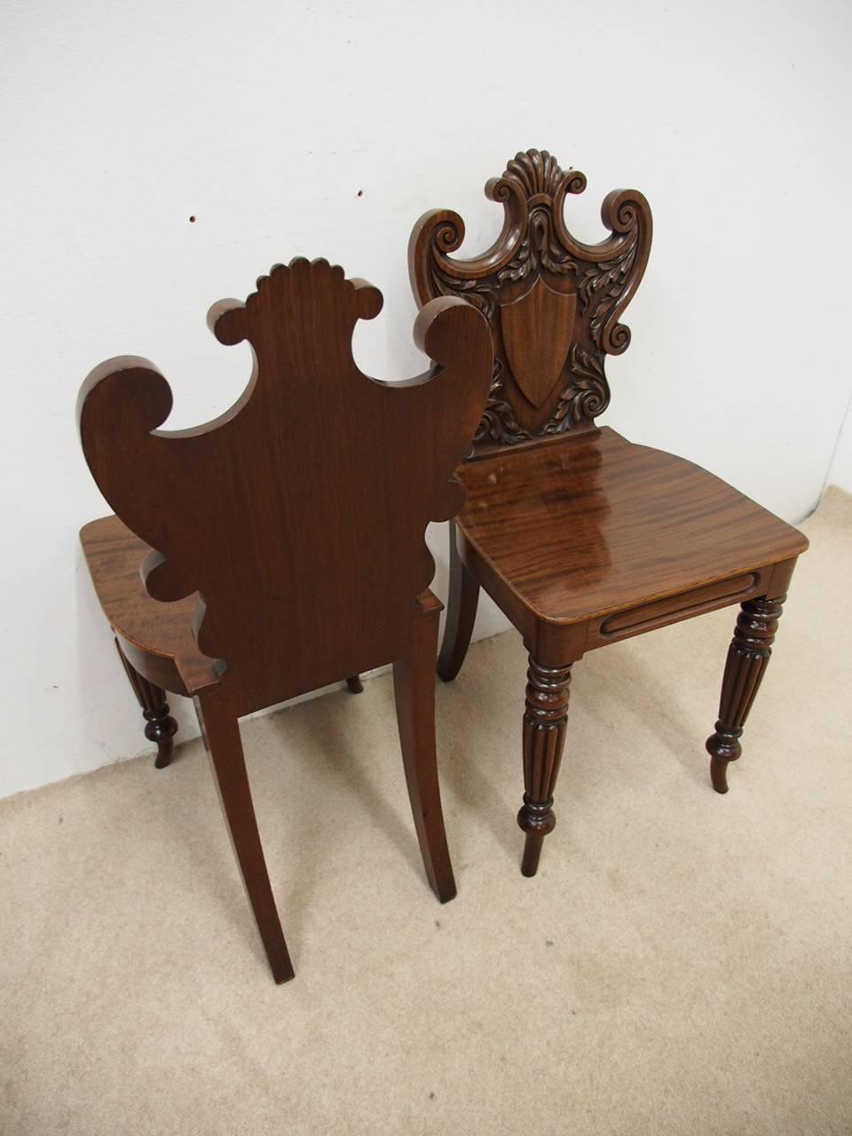 Exceptional pair of George III mahogany hall chairs, circa 1800. The elaborately carved shield-shaped backs are surmounted by a crest and with C-scroll carvings to the sides; a carved shield is centrally placed and surrounded by laurel leaves. The