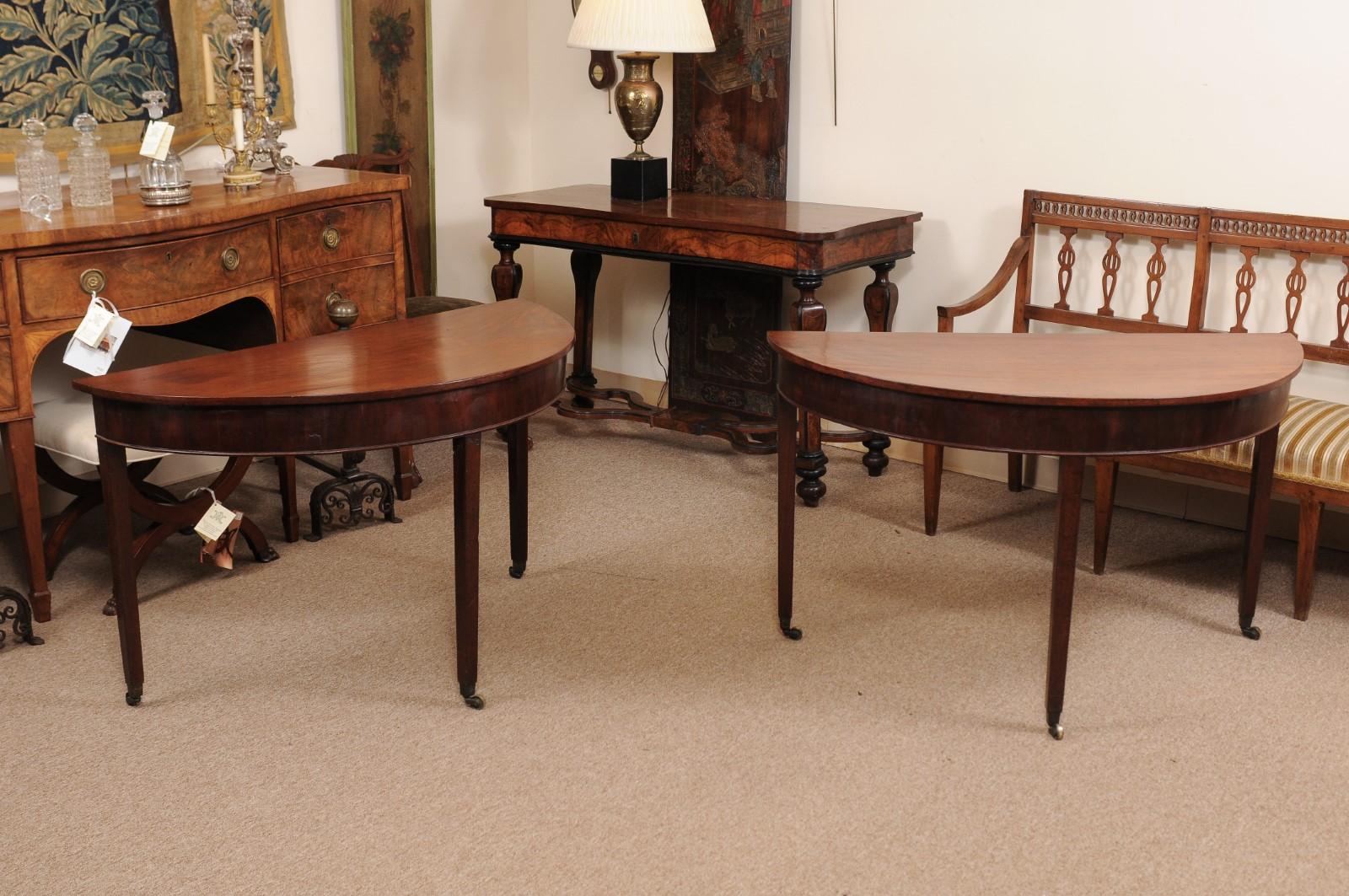 The pair of George III English mahogany demilune console tables with tapered legs and brass caster feet. 

 